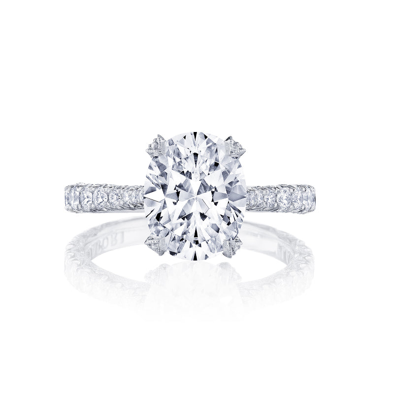 The Tacori Petite Crescent RoyalT Platinum Oval Solitaire Semi Mount engagement ring showcases a sparkling oval diamond on a reflective surface, complemented by a diamond-studded band featuring smaller round diamonds.
