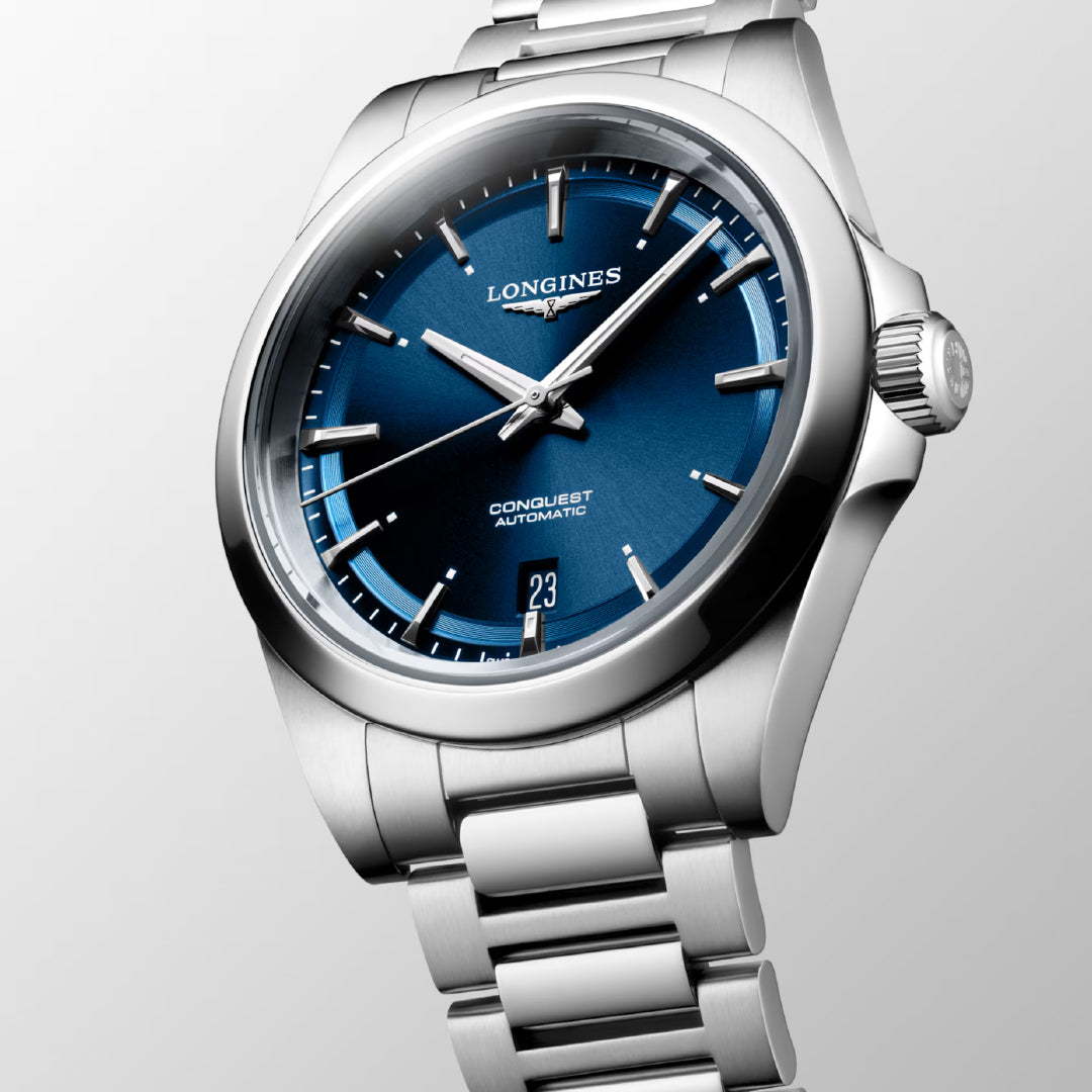 The Longines Conquest Automatic 38mm Watch features a blue dial, silver hour markers, and a date display at 6 o’clock, with a self-winding mechanical movement on a stainless steel bracelet.
