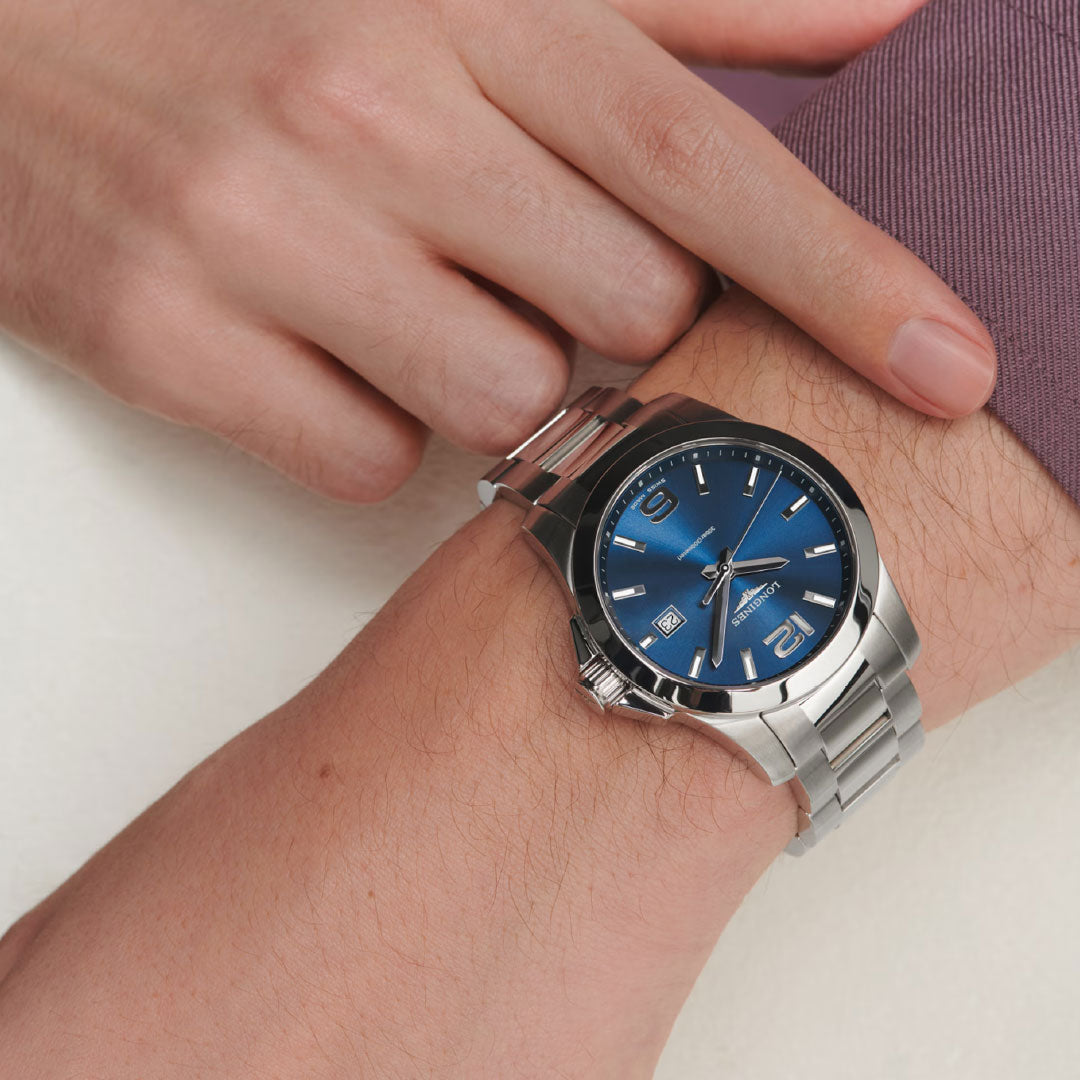 A person in a long-sleeved purple shirt adjusts the clasp of a Longines Conquest Quartz 41mm watch with a blue dial, highlighting sporty elegance.