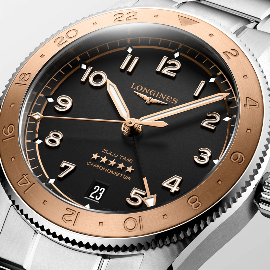 A close-up of the Longines Spirit Zulu Time 1925 Automatic 39mm Watch with a black dial, gold bezel, silver band, gold Arabic numerals, three hands, date window, and "Zulu Time Chronometer" on the face.