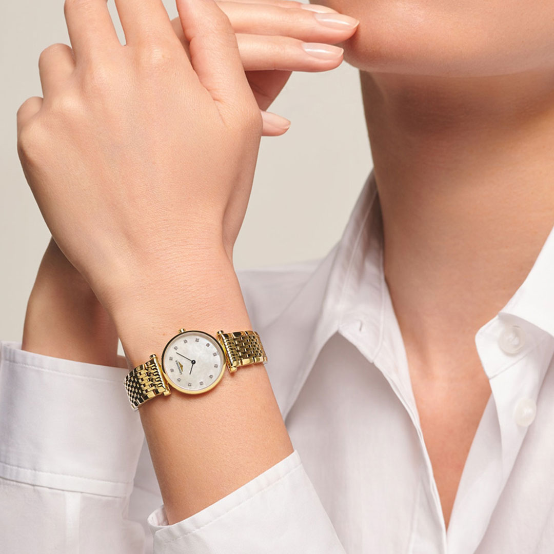 A person in a white button-up shirt rests their chin on one hand, highlighting the timeless style of the Longines La Grande Classique Quartz 24mm Watch with a white face and metal band. The background is plain and light-colored.