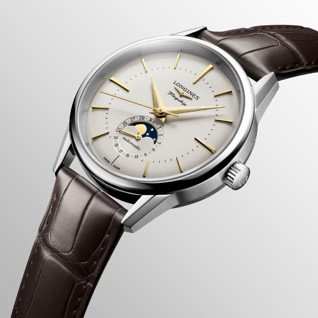 The Longines Flagship Heritage Moonphase Automatic 38.5mm Watch features a white dial, gold markers, moonphase subdial with moon and stars, and a brown leather strap, shown angled elegantly against a light gray background.