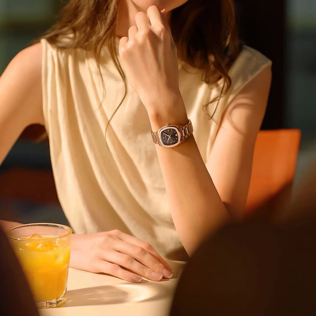 A person in a sleeveless beige top sits at a table, showcasing the MIDO Multifort TV Quartz 28mm Watch in silver. A glass of iced orange juice rests nearby on the table, while the individual’s face is partially out of frame.