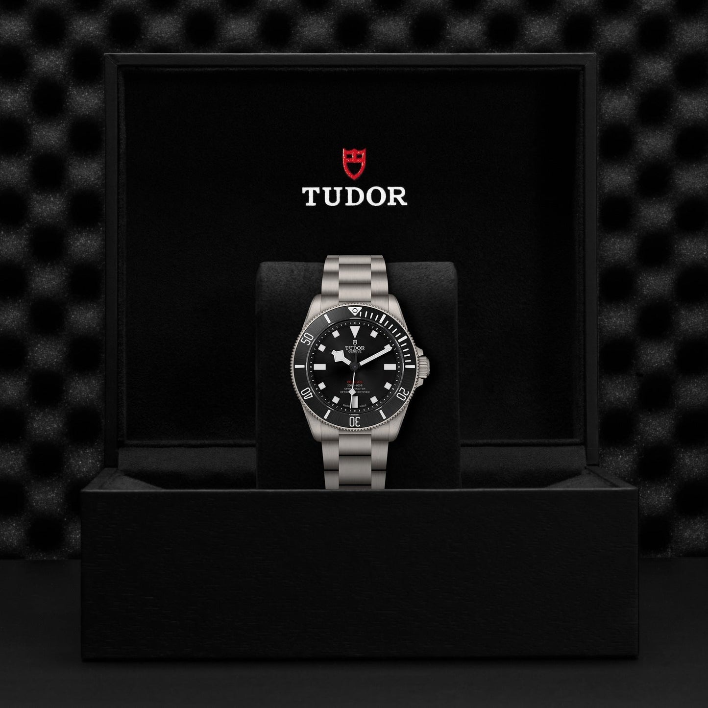 The TUDOR Pelagos 39, a luxury sports watch by TUDOR, features a sleek silver metal band and is showcased in an open black box. Its black watch face is adorned with white markers and a striking red logo, reminiscent of a dive watch, elegantly set against a textured black background.