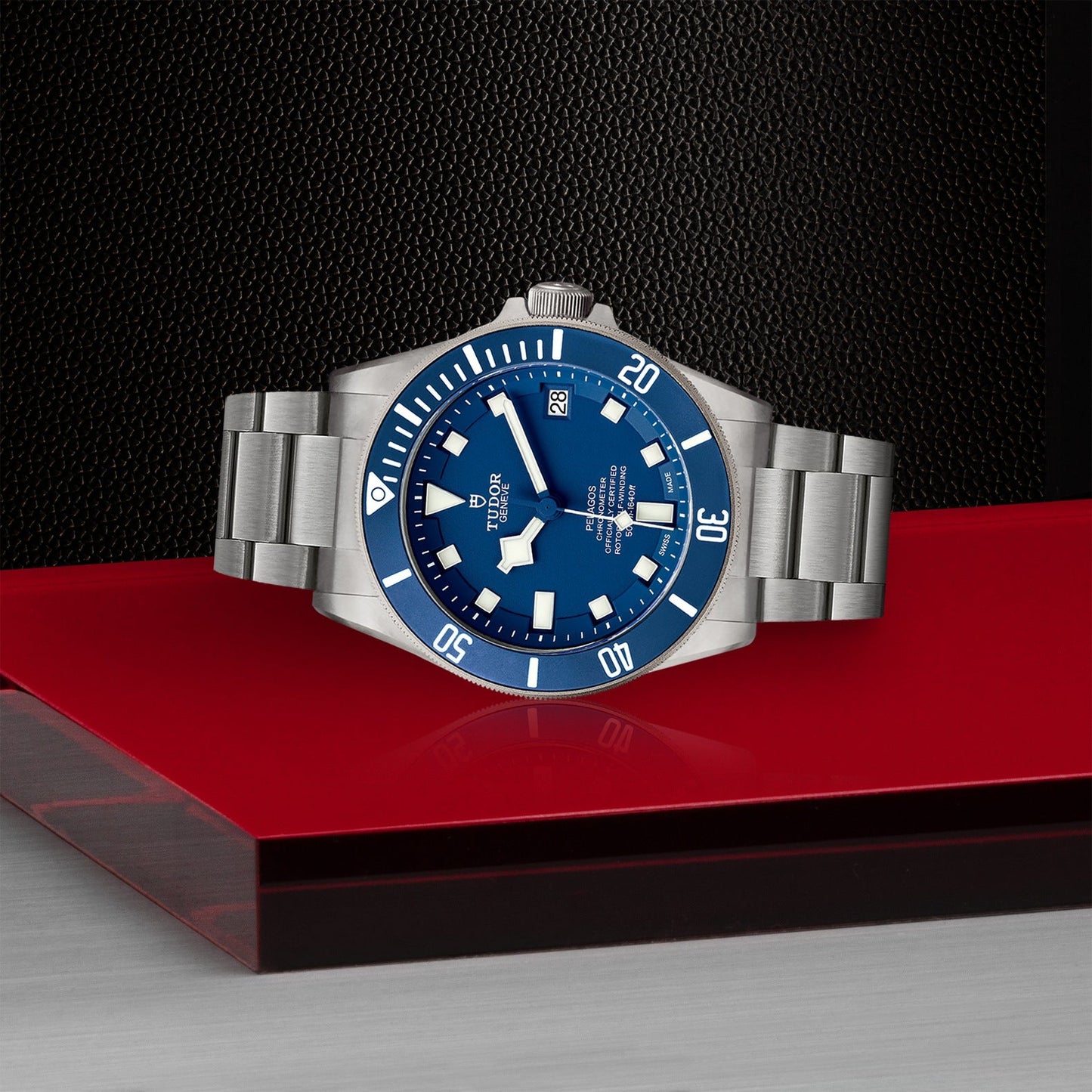 A TUDOR Pelagos stainless steel divers' watch, featuring a blue dial and rotating bezel, is showcased on a reflective red and gray surface. This TUDOR timepiece includes a date function, luminous markers, and boasts its waterproof design against a textured black background.