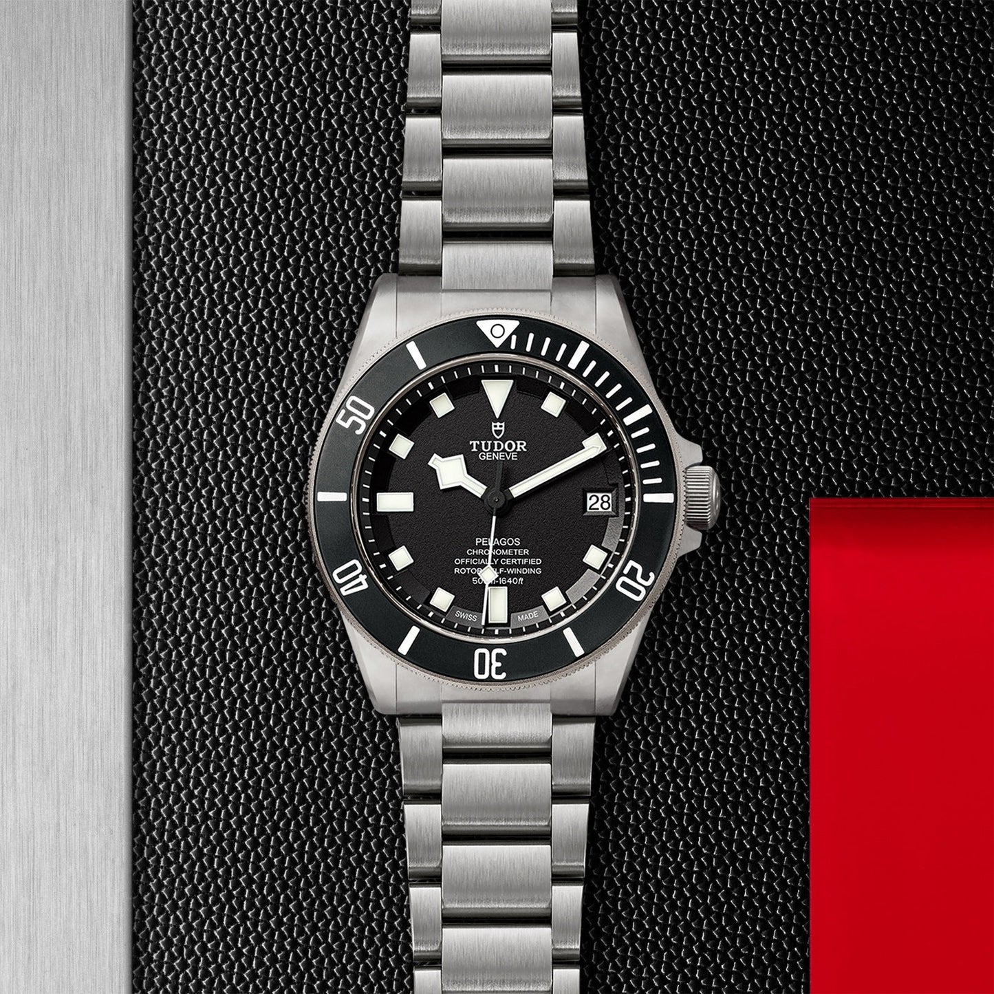 The TUDOR Pelagos, a silver wristwatch by TUDOR, is highlighted on a textured dark background with a red accent on the right. This remarkable timepiece features a black bezel and dial with luminous hour markers and a date display at 3 o'clock. It includes a titanium bracelet and is waterproof to 500m, making it perfect for diving enthusiasts.