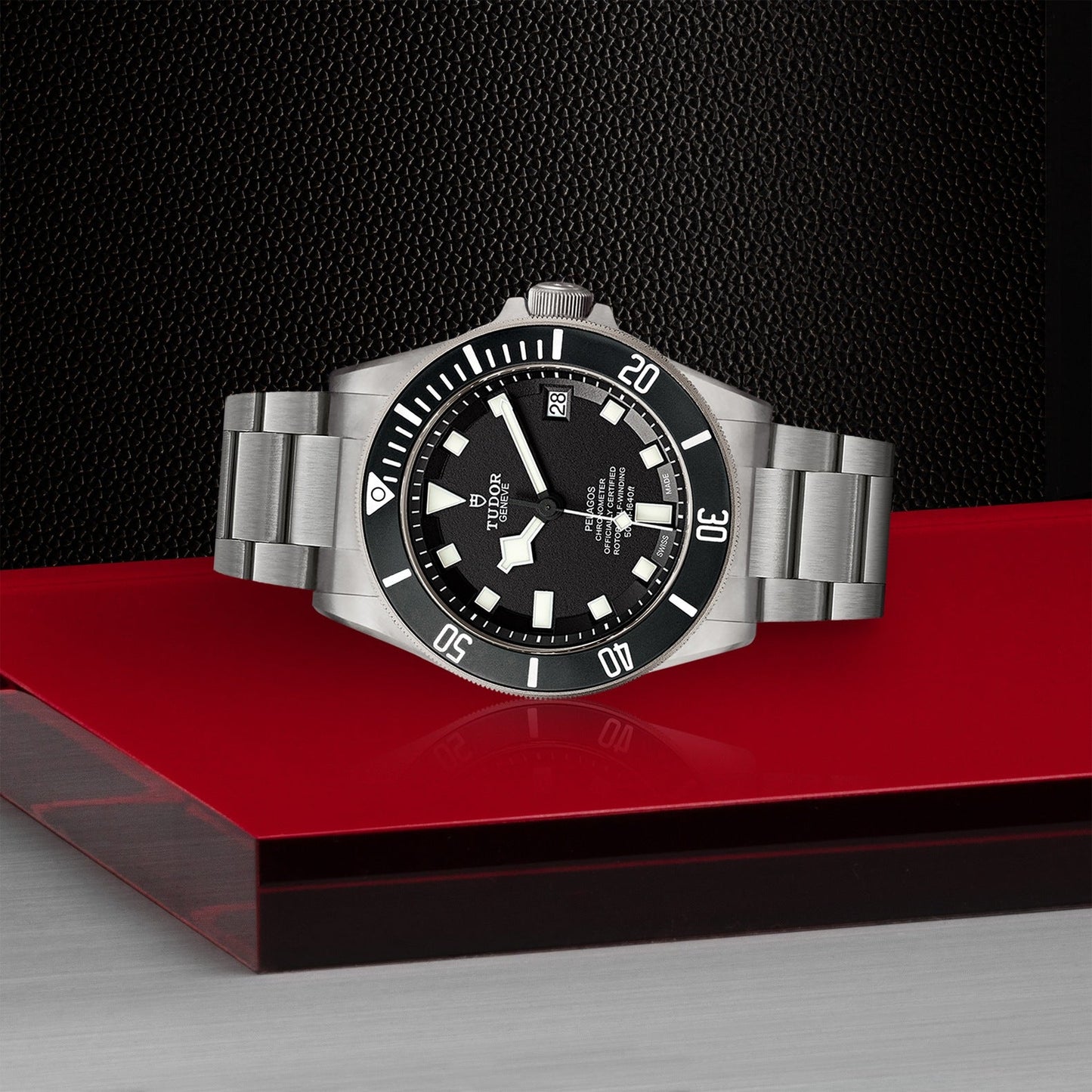 A TUDOR Pelagos, a luxury divers' watch with a stainless steel band and black dial, is displayed on a red and gray surface. The watch face features white markers, a date display, and proudly showcases the brand name "TUDOR." Perfect for aquatic adventures, it is waterproof up to 500m against a textured black background.