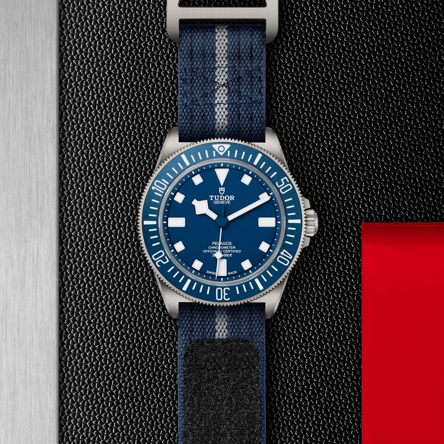 A close-up of the TUDOR Pelagos FXD wristwatch showcases its blue dial and bezel, complemented by a blue and gray fabric strap—highlighting the precision essential for professional use. The watch is set against a textured black background with a red rectangle on the right side.