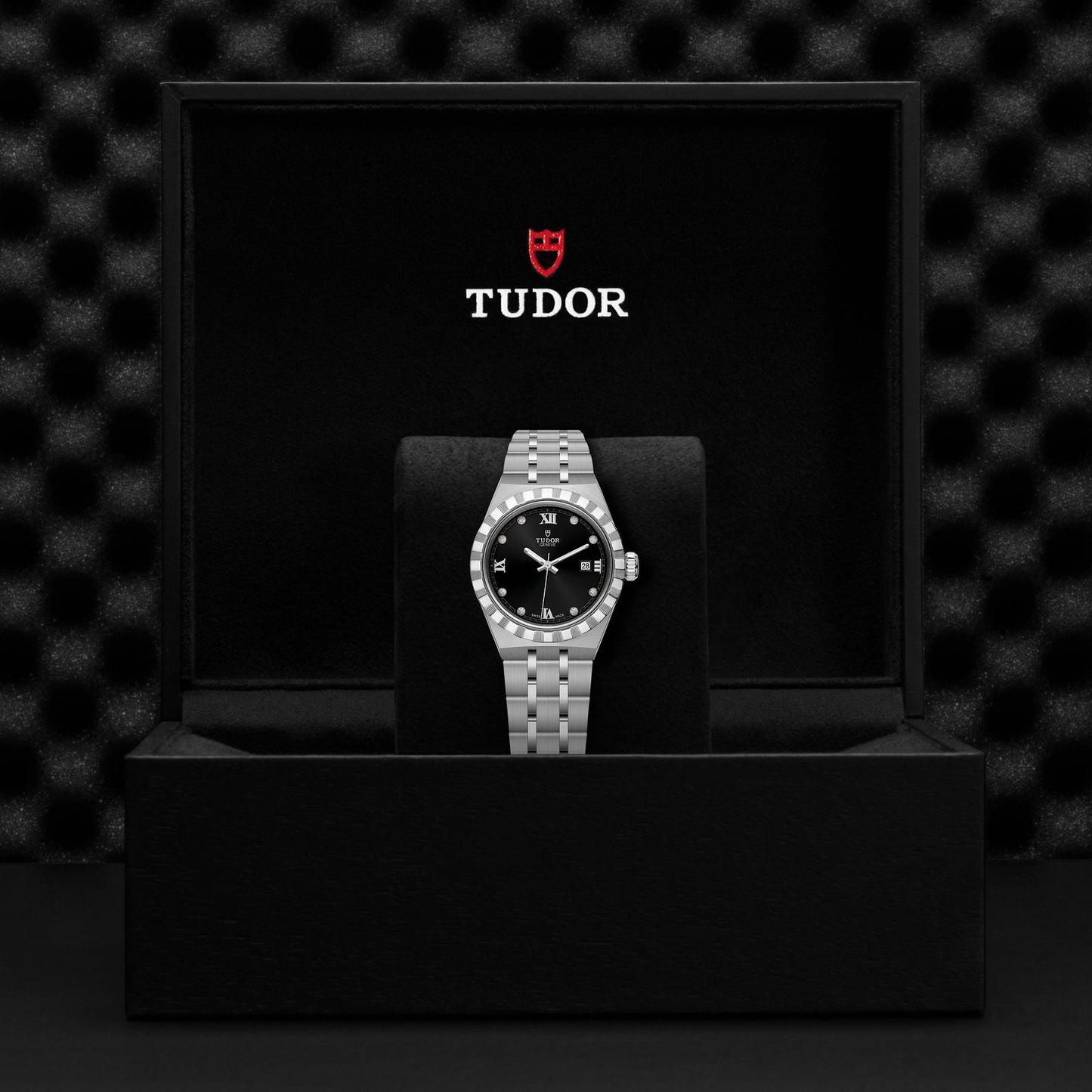 The TUDOR Royal watch, featuring a silver bracelet and a black dial adorned with diamonds, is showcased in an open black box. Inside the box, the striking red and white Tudor logo is prominently displayed against a textured dark background—exemplifying the sophisticated combination of an automatic sport-chic design.