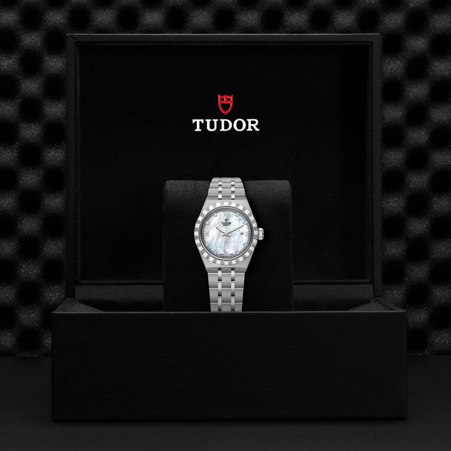 A TUDOR Royal watch, crafted from stainless steel with a silver finish, showcases its elegance with a metallic bracelet and diamond-studded bezel. It is beautifully presented inside an open black box featuring "TUDOR" in white, accompanied by a red shield logo on top. The dark, textured background accentuates its automatic sport-chic appeal.
