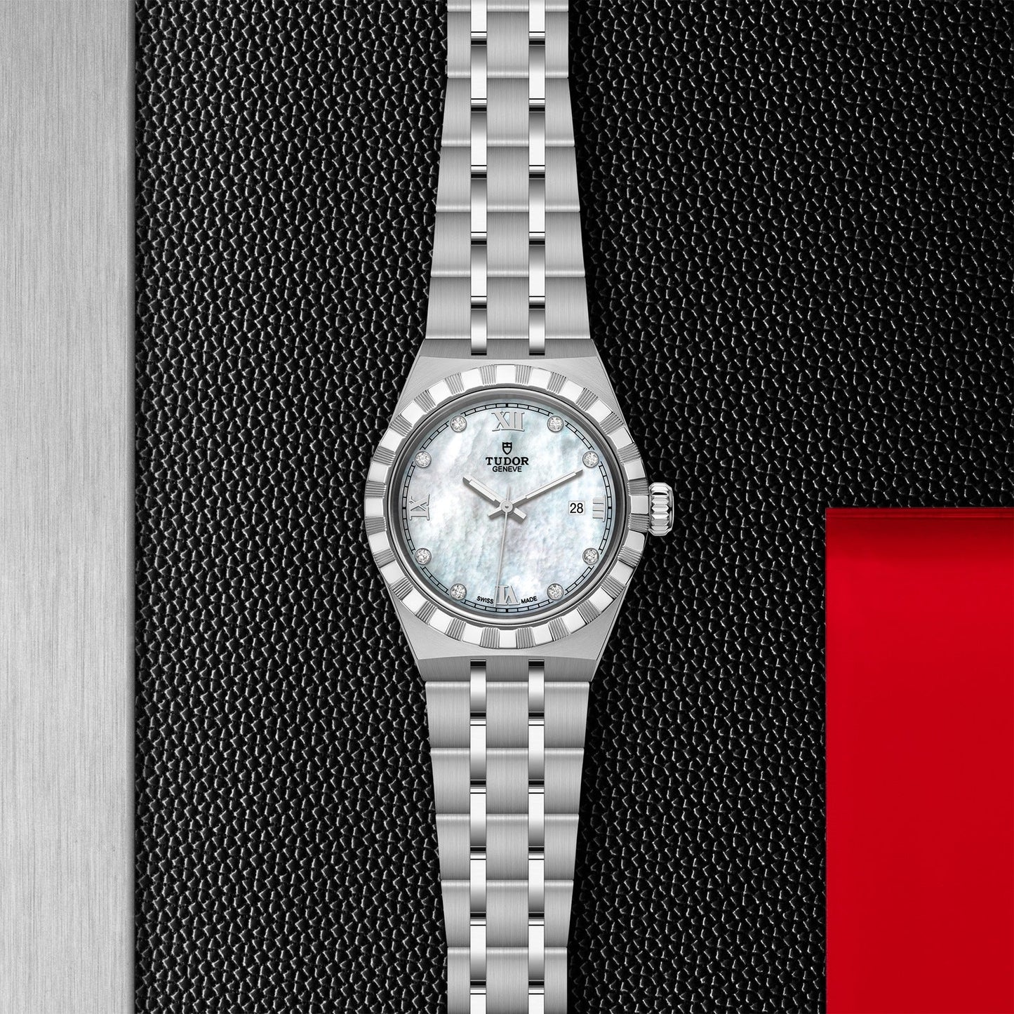 The TUDOR Royal wristwatch, featuring a white mother-of-pearl face with Roman numerals and a stainless steel link band, is elegantly showcased against a textured black background with hints of red. This model from TUDOR combines automatic sport-chic style with a convenient date window at 3 o'clock.