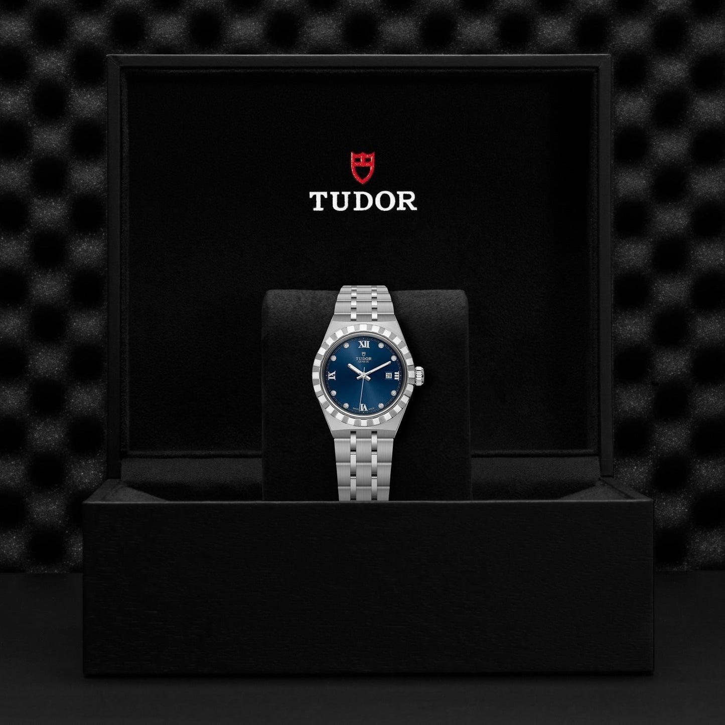 Inside an open black box, a TUDOR Royal wristwatch catches the eye with its stunning blue dial and date display. Crafted from stainless steel and gold, it exudes sport-chic elegance. The interior of the box is lined with black fabric, featuring a textured black backdrop that showcases the red TUDOR logo on the inside lid.