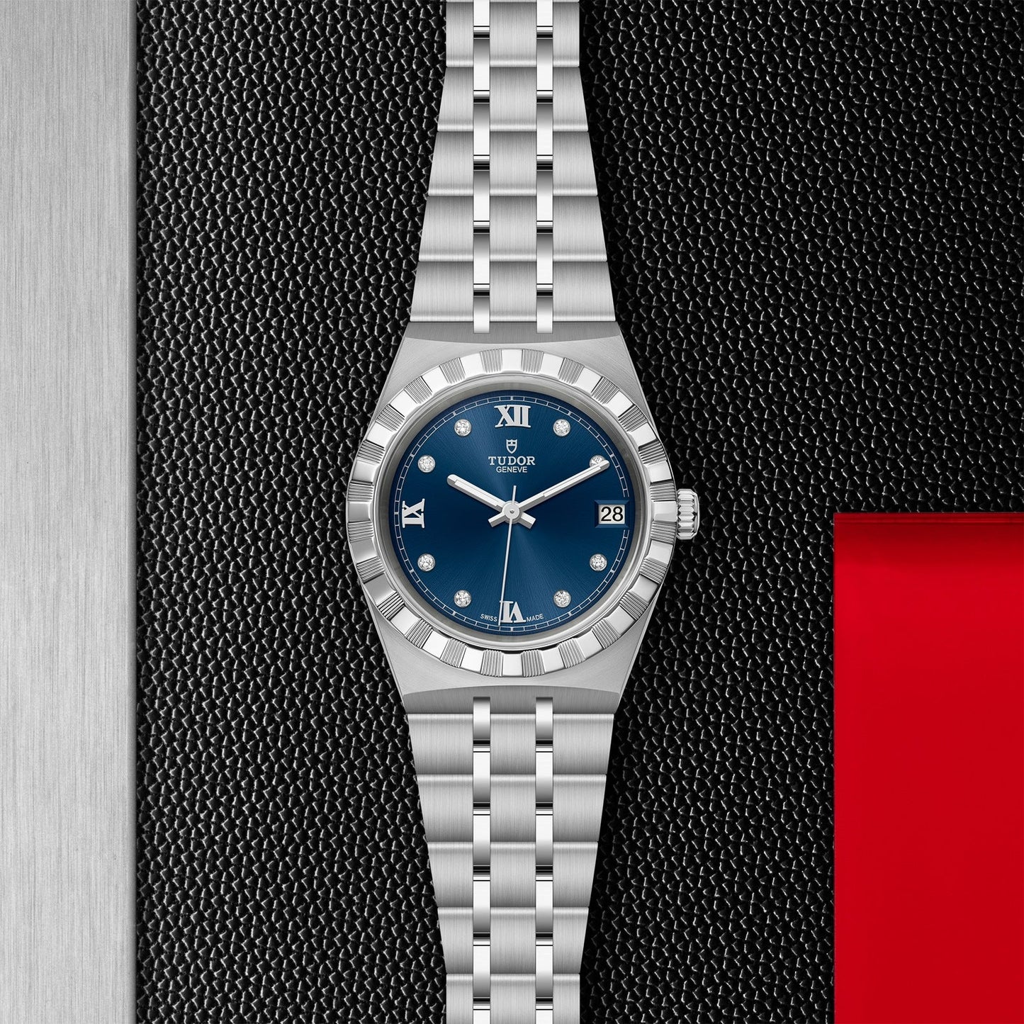 The TUDOR Royal watch by TUDOR, in a sophisticated silver finish, is beautifully showcased on a textured black surface. It features a blue dial adorned with diamonds, Roman numerals at 12 and 6 o'clock, a date window at 3 o'clock, and a distinctive red accent on the right. This model exemplifies the automatic sport-chic style.