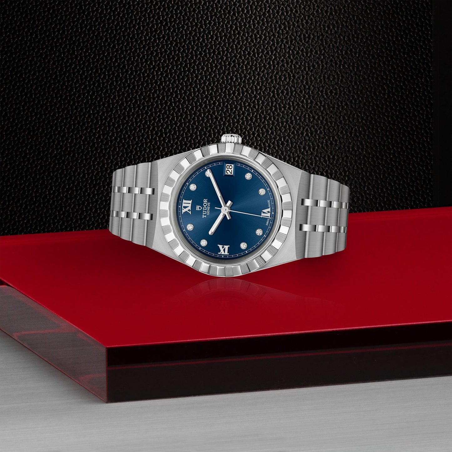 A TUDOR Royal luxury watch showcases a silver design with a blue dial adorned by Roman numeral markers, elegantly set against a glossy red and gray surface with a textured black background. This sophisticated timepiece is equipped with a metal bracelet and date window, perfectly embodying the automatic sport-chic aesthetic.