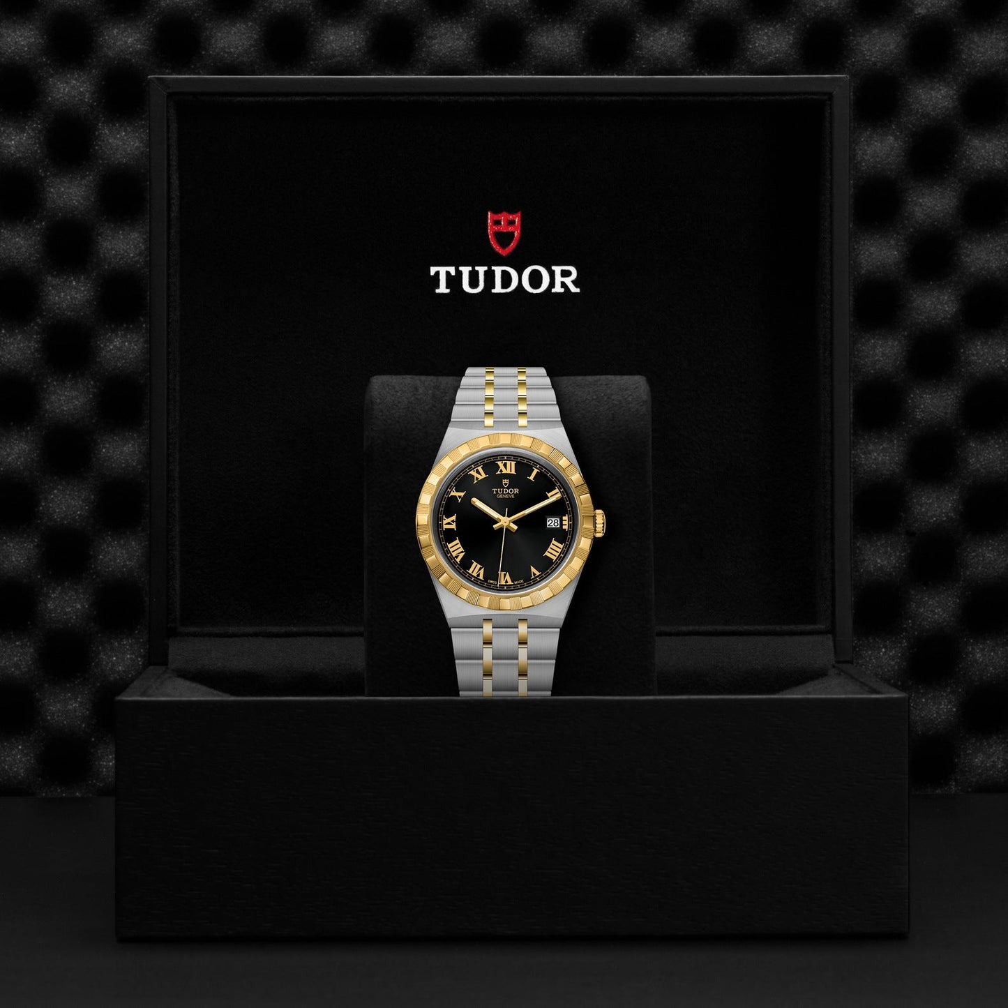 A TUDOR Royal wristwatch is presented in a stylish black box, featuring an automatic sport-chic design with a gold-accented integrated bracelet and a black face adorned with gold Roman numerals. The elegant display includes the TUDOR Royal logo inside the box.