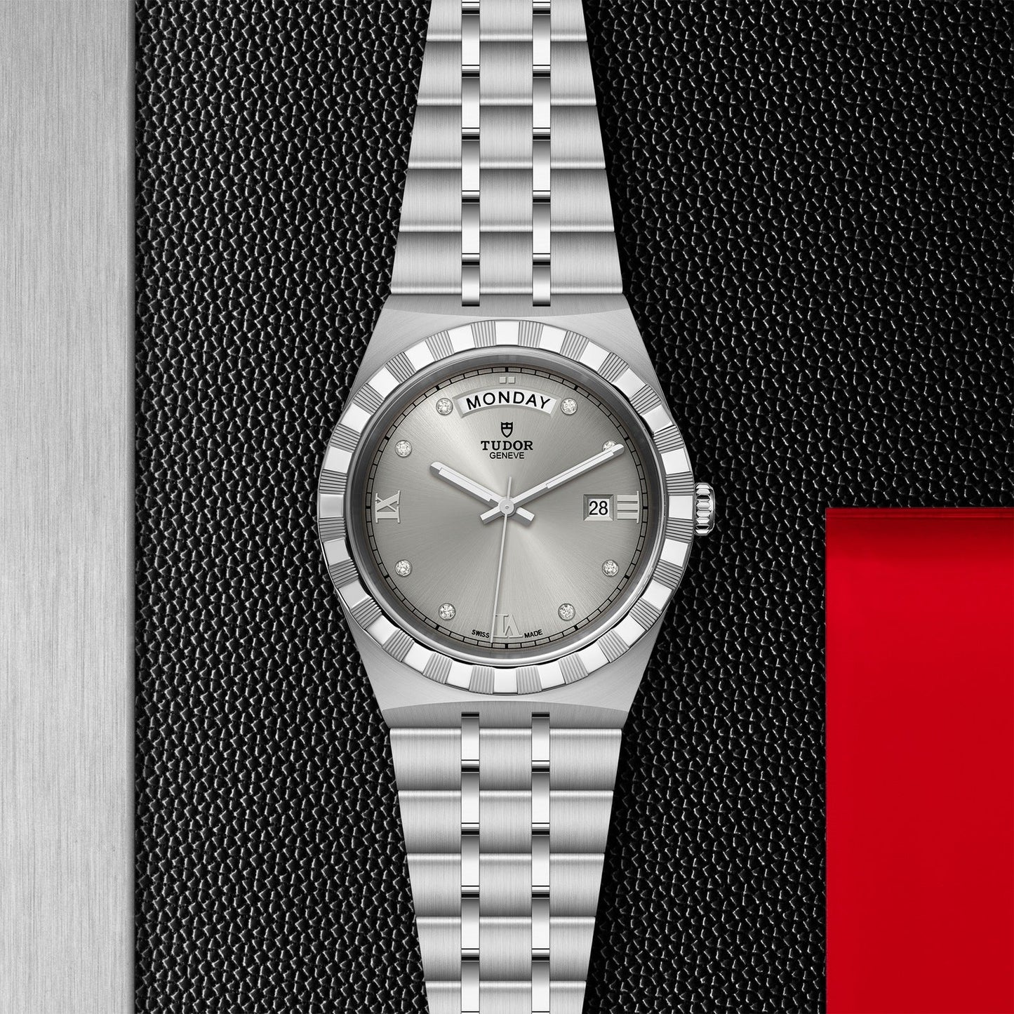 Displayed against a textured black background, the TUDOR Royal wristwatch from TUDOR features a silver round face and a metal bracelet. The watch showcases automatic sport-chic elegance with the day "Monday" and date "28" displayed, while its sides are framed by red and metallic elements.