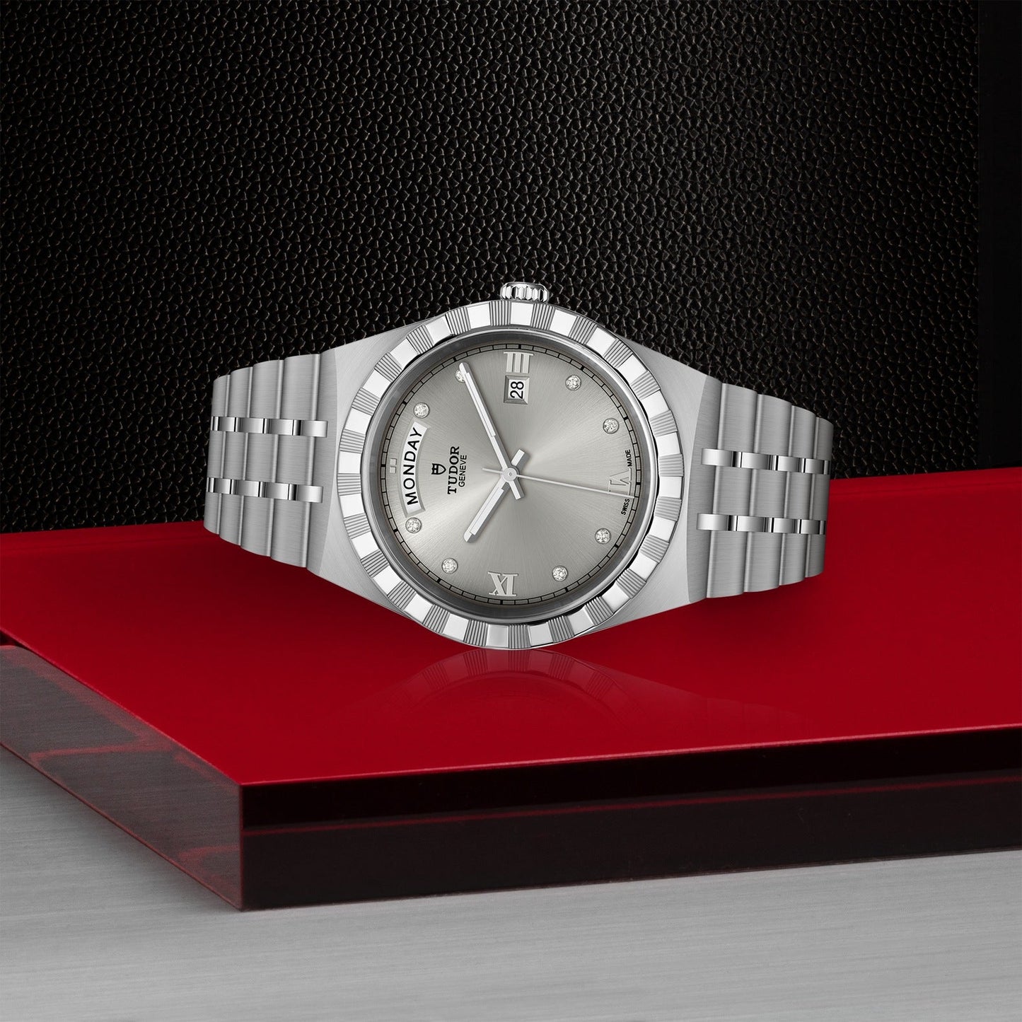 A TUDOR Royal wristwatch in silver, featuring a metallic strap, is showcased on a red and black platform. The watch's face includes Roman numerals, a day and date display, along with a textured bezel. This sport-chic automatic timepiece by TUDOR stands out against the black textured pattern background.