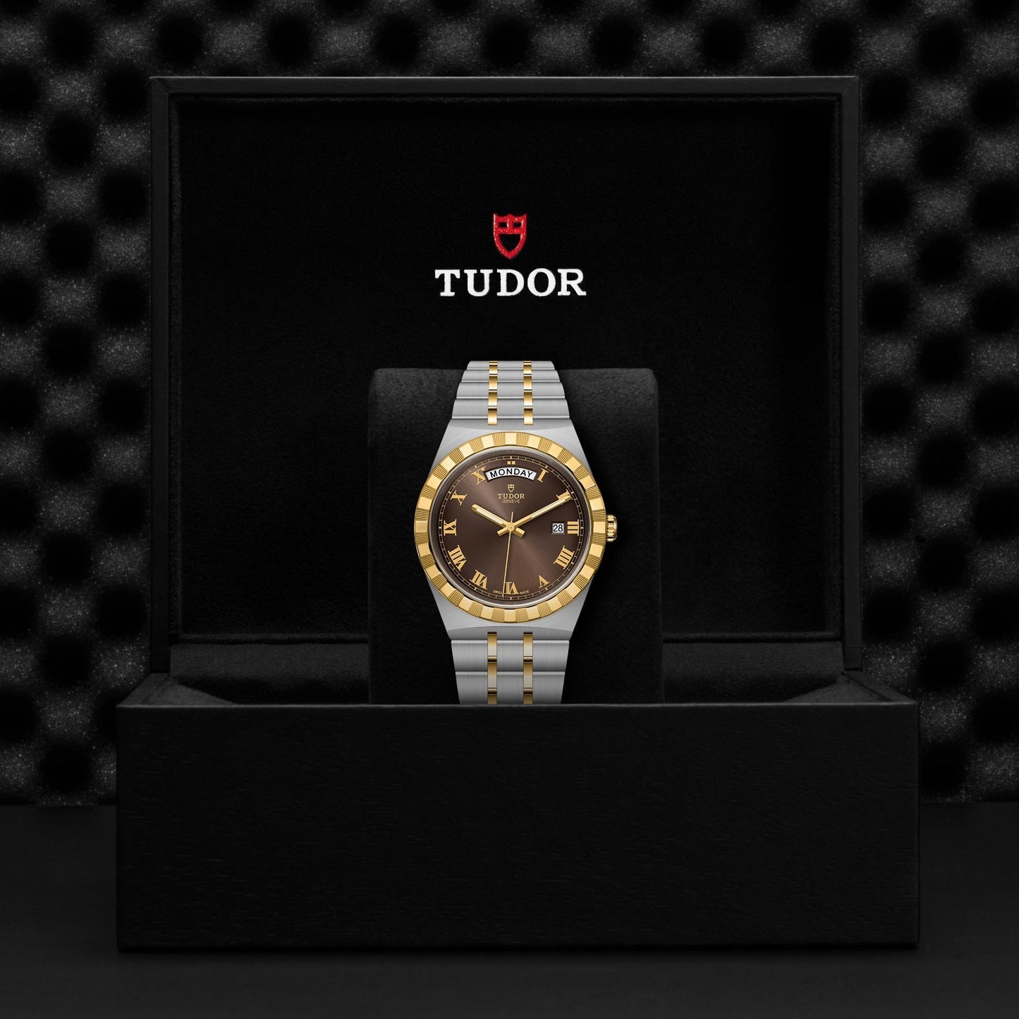 Displayed inside a black box is a TUDOR Royal wristwatch, crafted with a stainless steel and gold two-tone band. This automatic sport-chic timepiece showcases a brown dial featuring Roman numeral hour markers and is complemented by a gold-accented bezel. The esteemed Tudor logo sits proudly above this elegant watch inside the box.