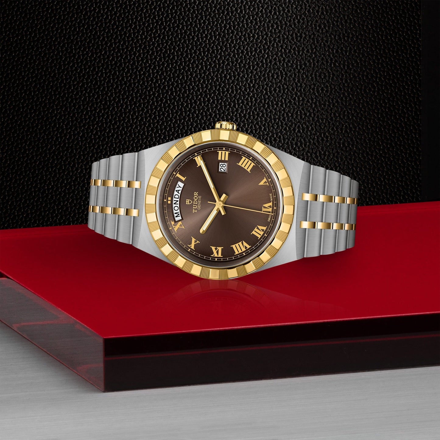 The sophisticated TUDOR Royal wristwatch showcases a brown and black dial adorned with gold Roman numerals and a day/date display. This automatic sport-chic timepiece features a stainless steel and gold band, elegantly positioned on a red and black surface with a textured black background.