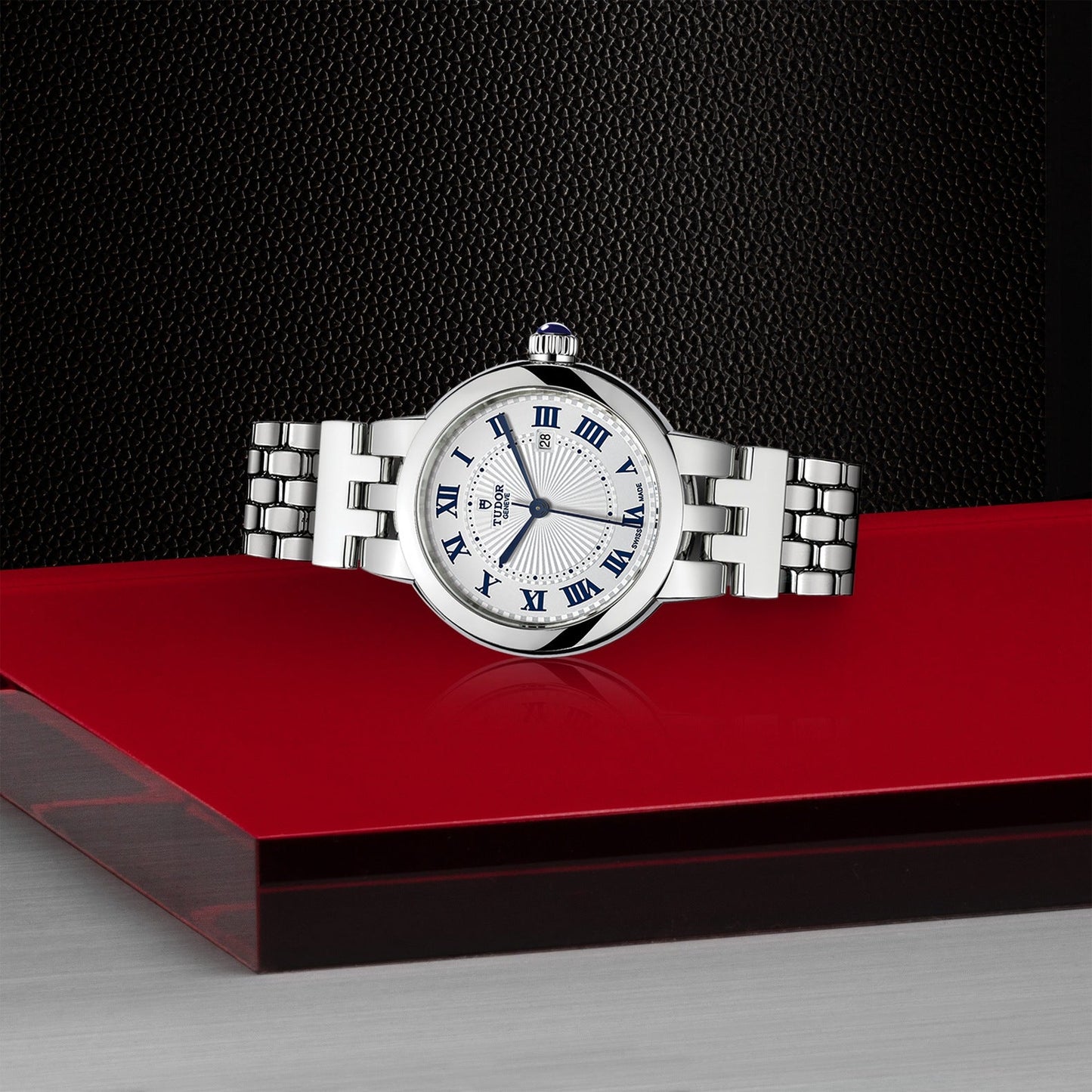 The TUDOR Clair de Rose wristwatch features an opaline dial with Roman numerals and a striped center pattern. Elegantly displayed on a glossy red surface against a textured black backdrop, it exudes timeless charm with its classic design.