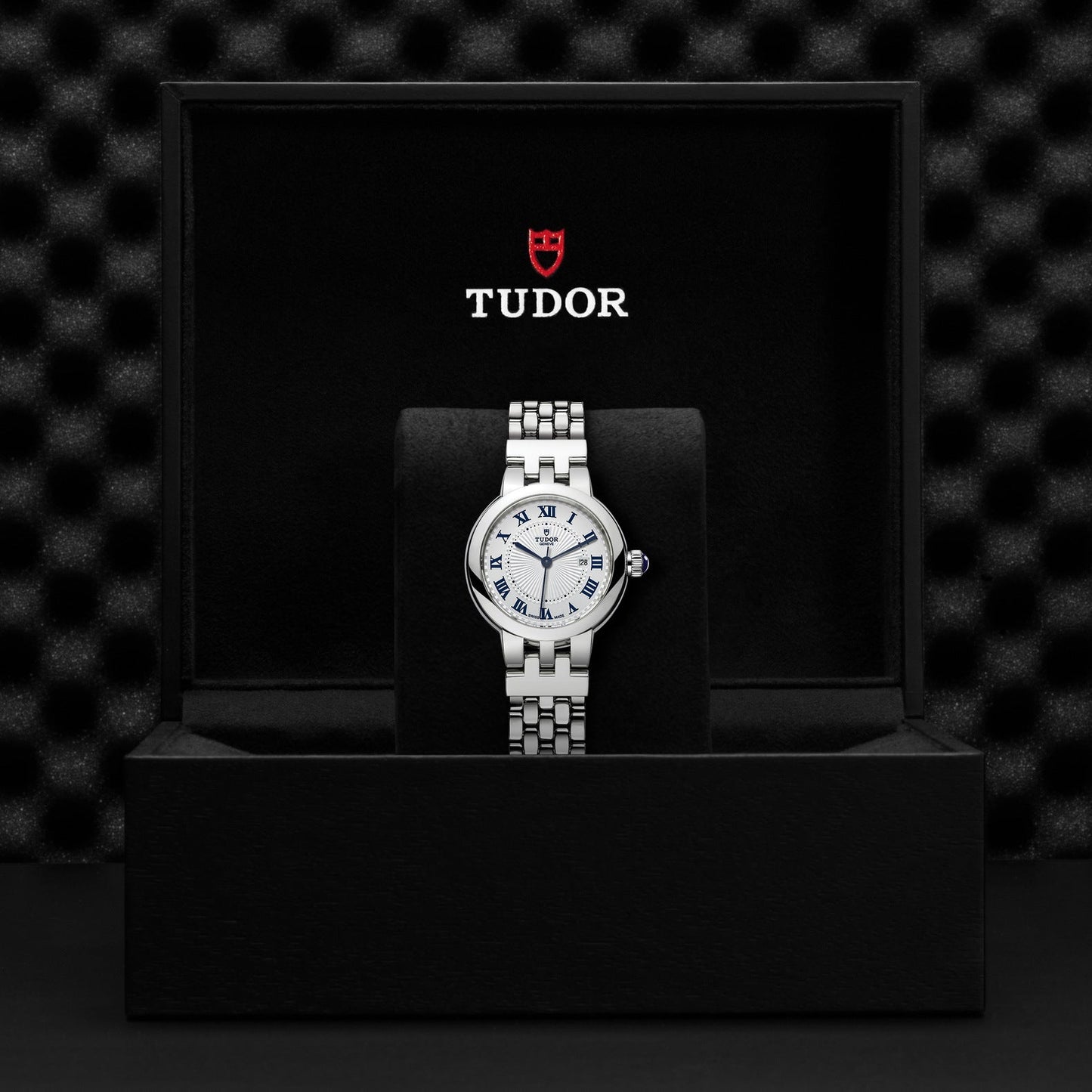 A TUDOR Clair de Rose wristwatch, featuring an opaline dial adorned with Roman numerals, is showcased in an open black box. The TUDOR logo is prominently displayed inside the lid against a textured black background.
