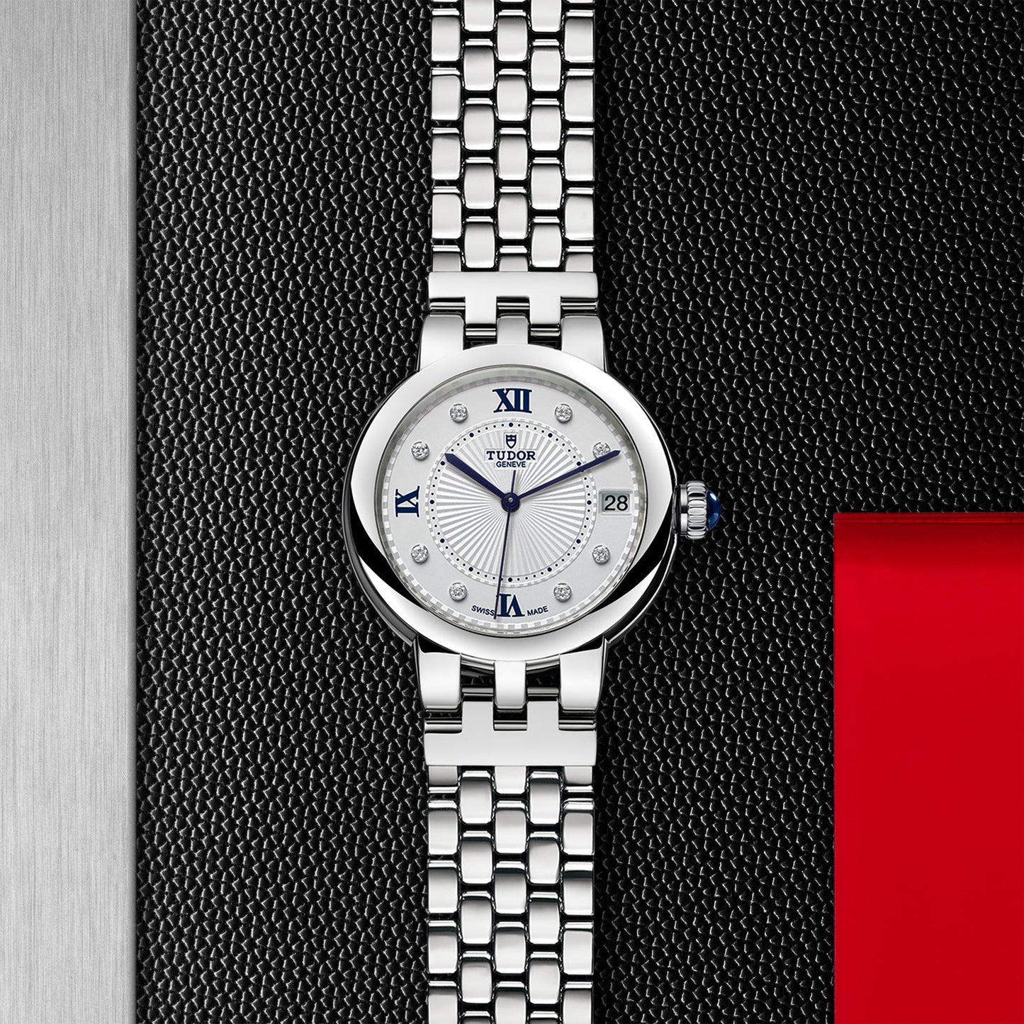 The TUDOR Clair de Rose wristwatch features a silver finish with a patterned metal band and an opaline dial. It elegantly displays Roman numerals and diamond hour markers, set against a backdrop that includes a black textured surface, a red square, and a metallic vertical strip, embodying the sophisticated TUDOR style.