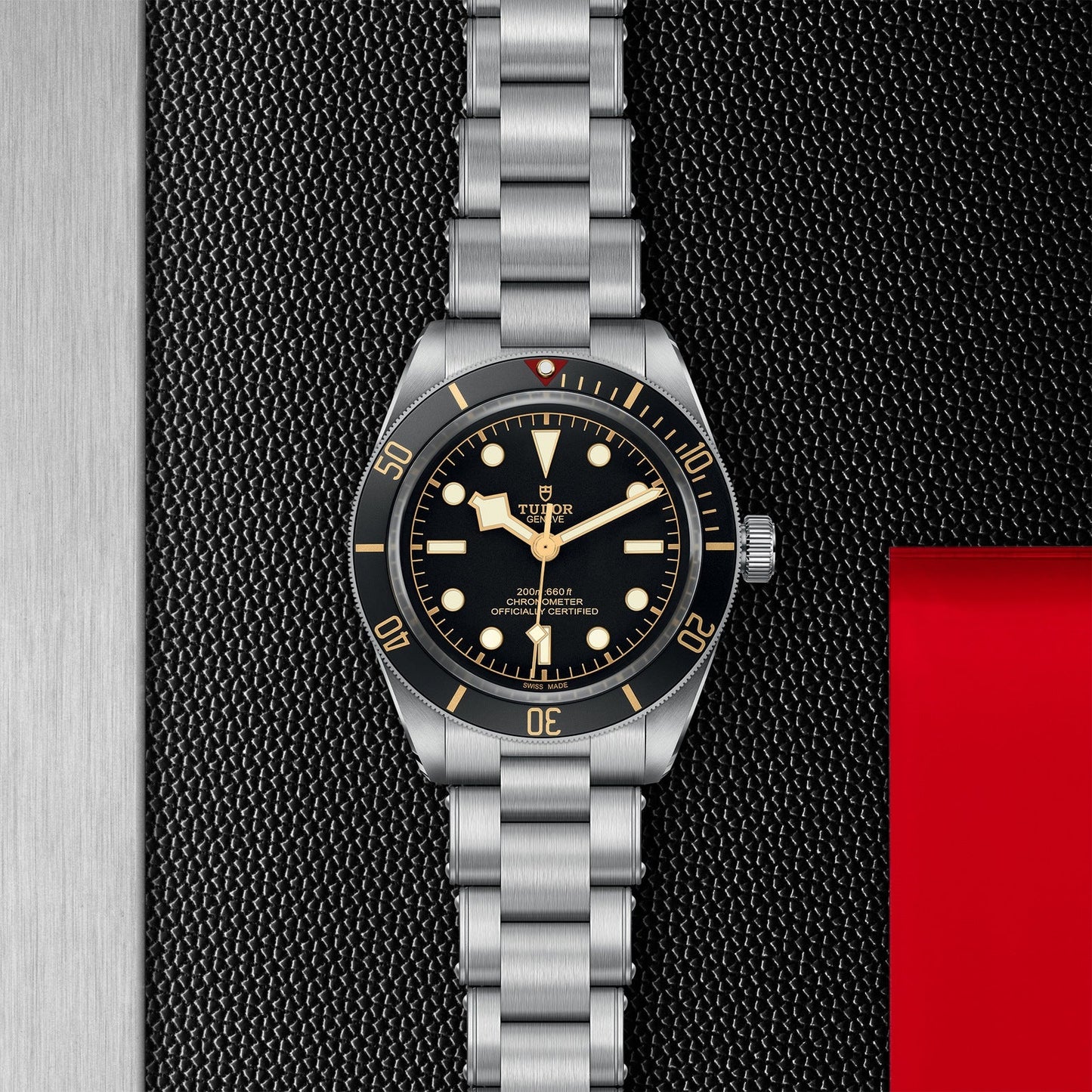 The TUDOR Black Bay 58 is a stylish dive watch featuring a sleek silver wristwatch design with a black bezel and face. It includes luminous hour markers and hands, set against a textured black background, completed with a distinctive red accent on the right side, all paired with a steel bracelet.