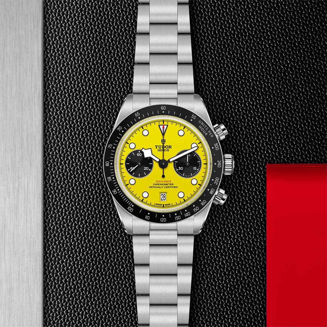 A TUDOR Black Bay Chrono 39 stainless steel wristwatch with a bright yellow dial, black subdials, and metal bracelet is displayed on a textured black surface accented with red and metallic silver strips.