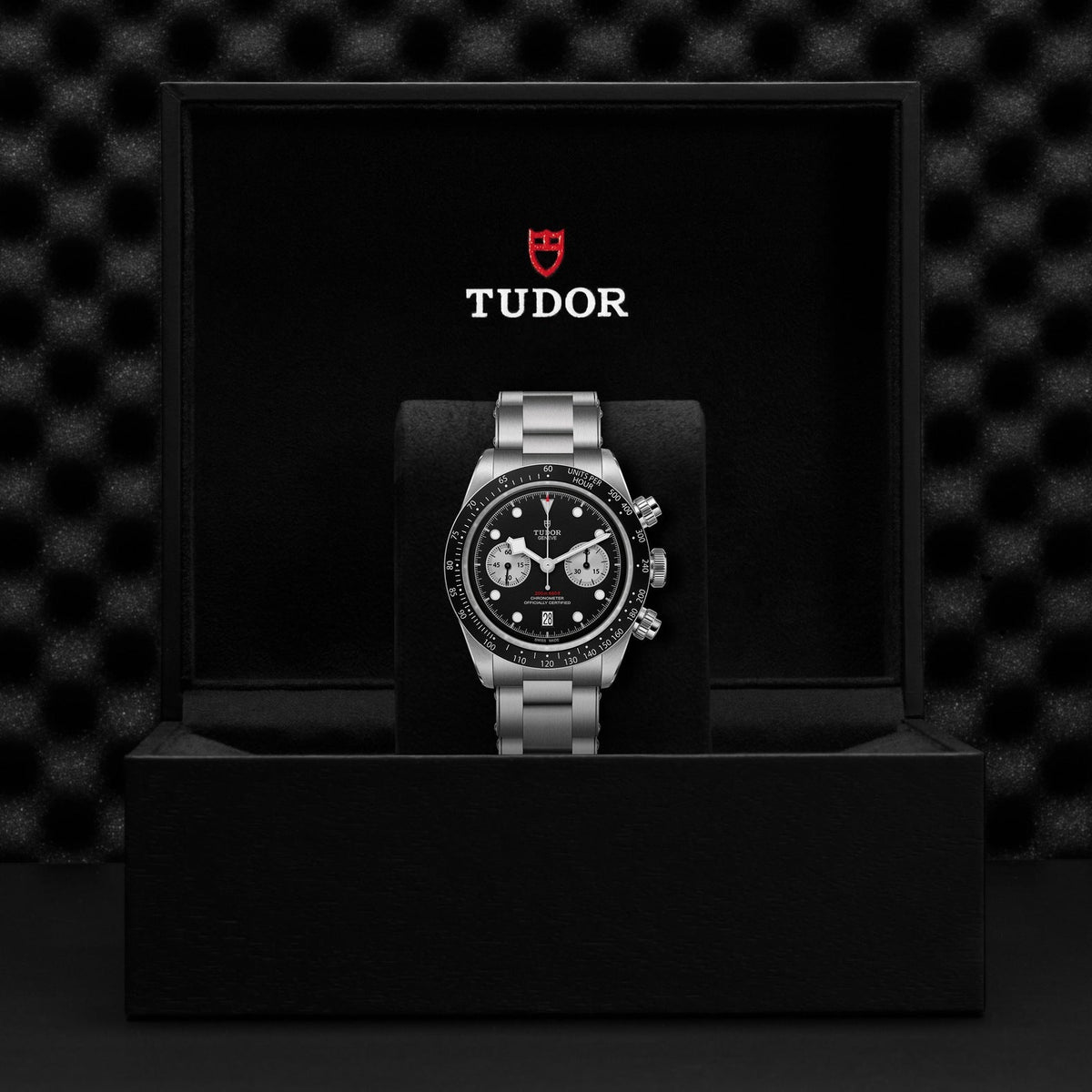 The TUDOR Black Bay Chrono boasts a stainless steel band and an eye-catching black dial, showcased in an open black case. Inside, the box is elegantly lined with smooth black fabric, featuring the TUDOR logo prominently displayed in white and red. A textured dark surface behind it adds to its sophistication.