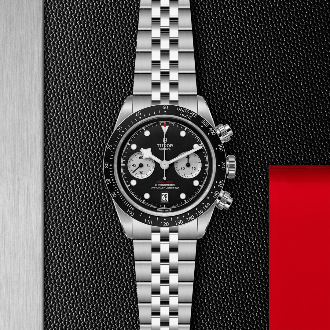 Discover the TUDOR Black Bay Chrono, a stylish sports watch with a steel case, black dial, three sub-dials, date display, and tachymetric scale bezel. It includes a link bracelet against a textured black backdrop with red and gray accents.
