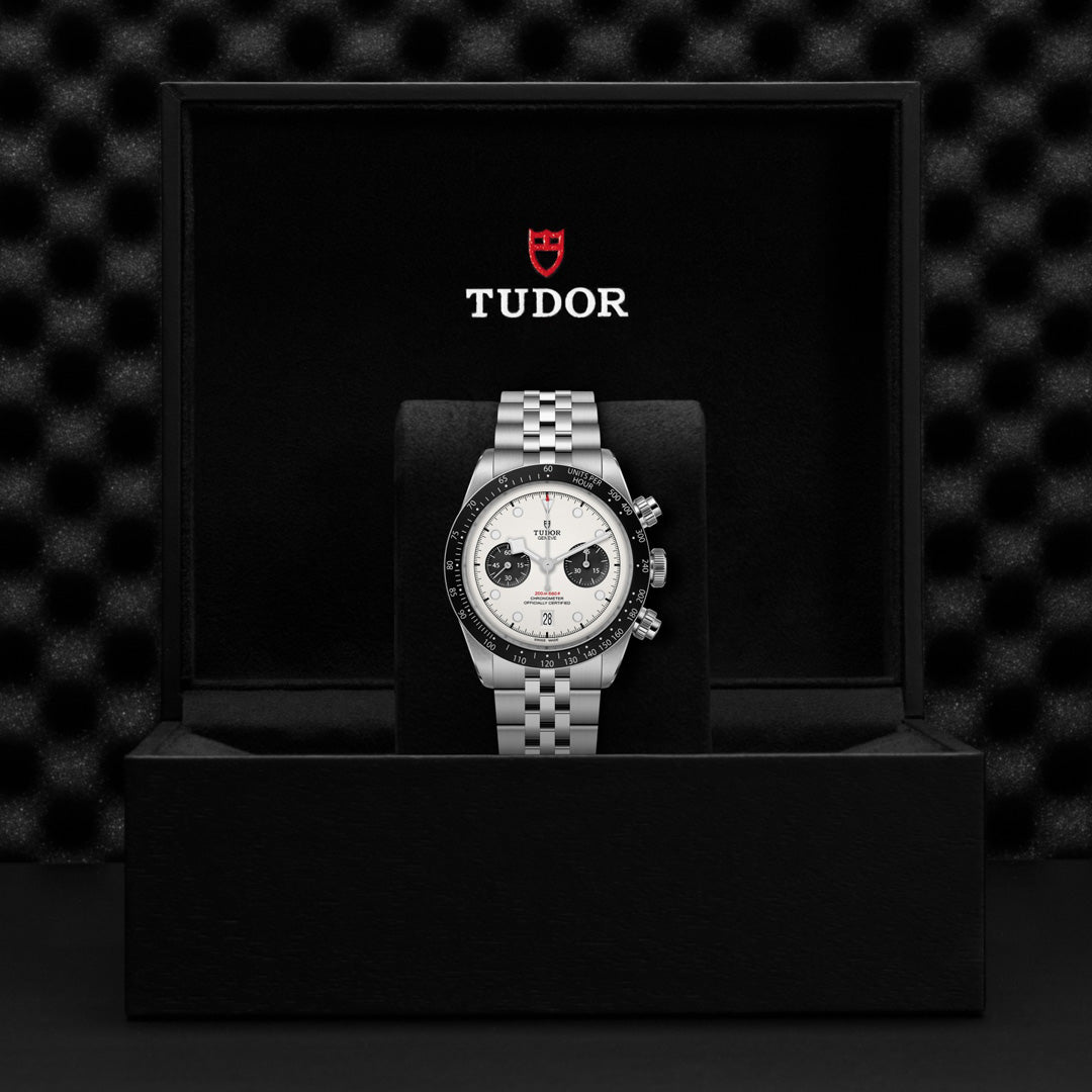 The TUDOR Black Bay Chrono, a luxury wristwatch with a silver band and dual-tone dial, is stylishly presented in an open black box. Inside the lid, the "TUDOR" logo is prominently featured in white and red against a textured dark background.