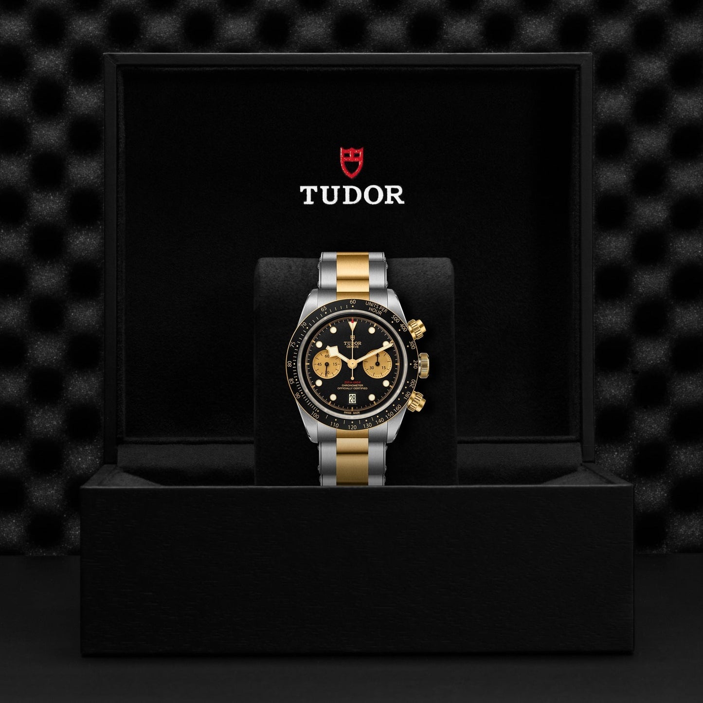 The TUDOR Black Bay Chrono S&G watch comes packaged in a sleek black presentation box with a textured interior. This chronograph boasts a black dial complemented by gold subdials and a two-tone gold and silver strap, with the TUDOR logo elegantly displayed inside the lid.