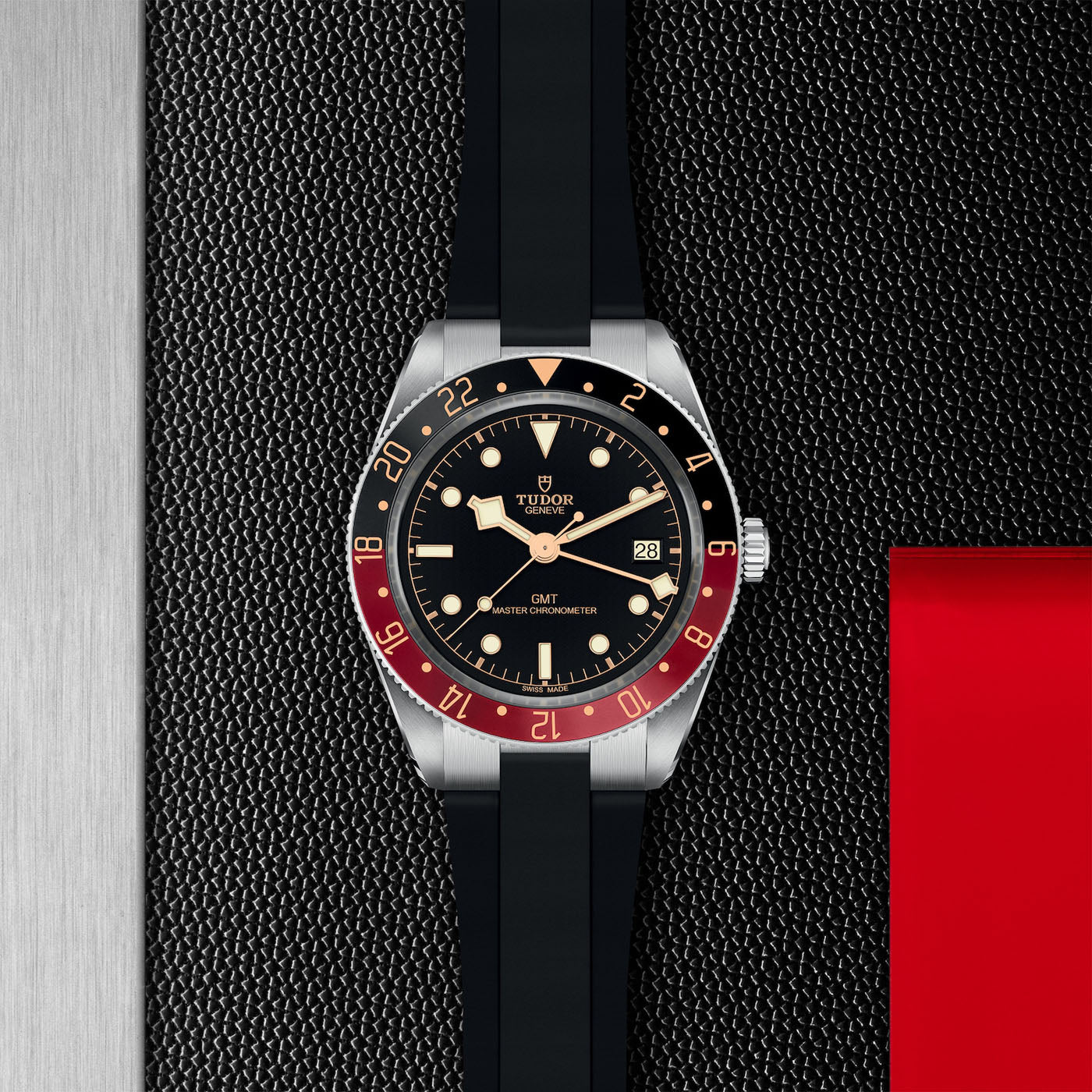 The TUDOR Black Bay 58 GMT dive watch features a black dial, a red and black rotating bezel, and a robust black rubber strap. Displayed against a textured dark background with an eye-catching red stripe, it includes silver hour markers and a date window positioned at 3 o'clock.