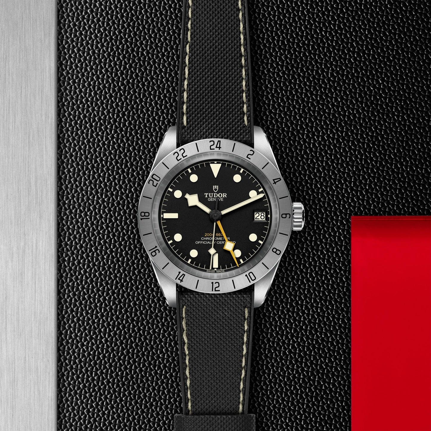 The TUDOR Black Bay Pro wristwatch features a black dial and silver bezel, elegantly placed on a dark textured surface next to a metallic panel with a red square accent. It comes equipped with luminescent markers, dual time zone functionality, the TUDOR Manufacture Calibre MT5652 movement, date display, and is paired with a black strap featuring white stitching.