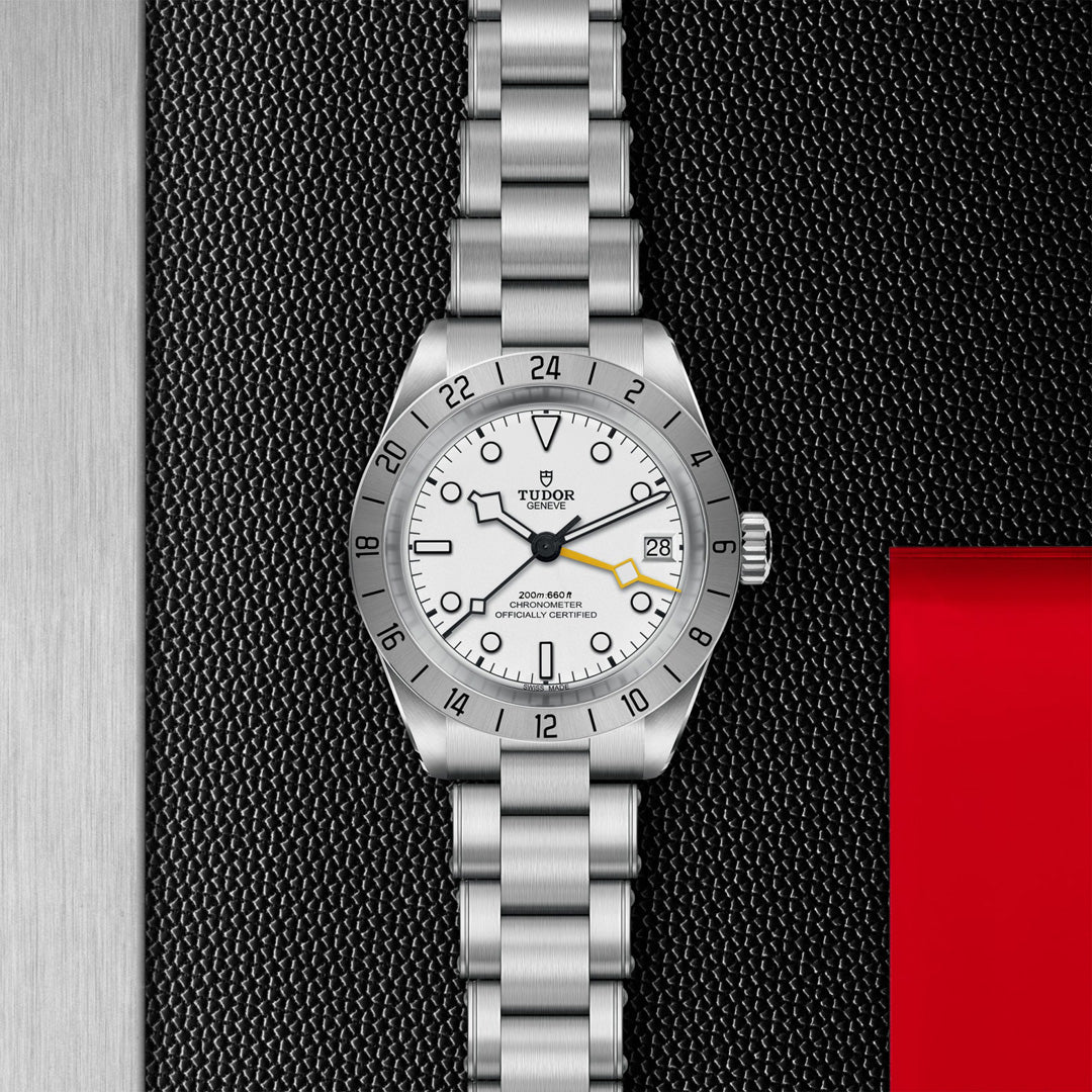 The TUDOR Black Bay Pro is a stainless steel watch with a white dial, silver hour markers, and date window. It features the TUDOR Manufacture Calibre MT5652 with dual time zone capabilities, showcased on a textured black and red background.