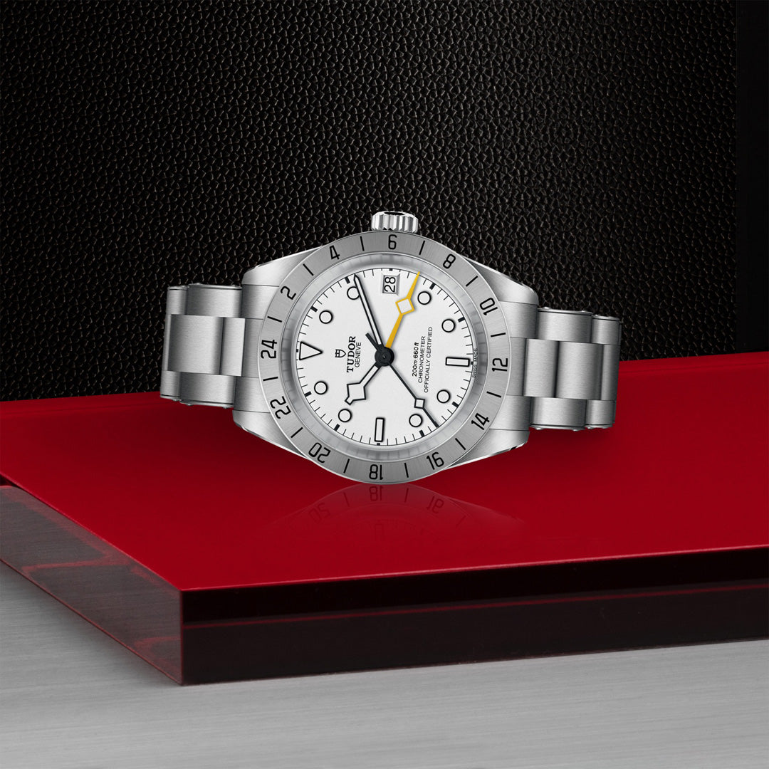 The TUDOR Black Bay Pro is elegantly displayed at an angle, highlighting its silver stainless steel build and TUDOR Manufacture Calibre MT5652. It features a white face with black numerals, a yellow-tipped hand, and a date function on a red and black platform set against a textured black background.