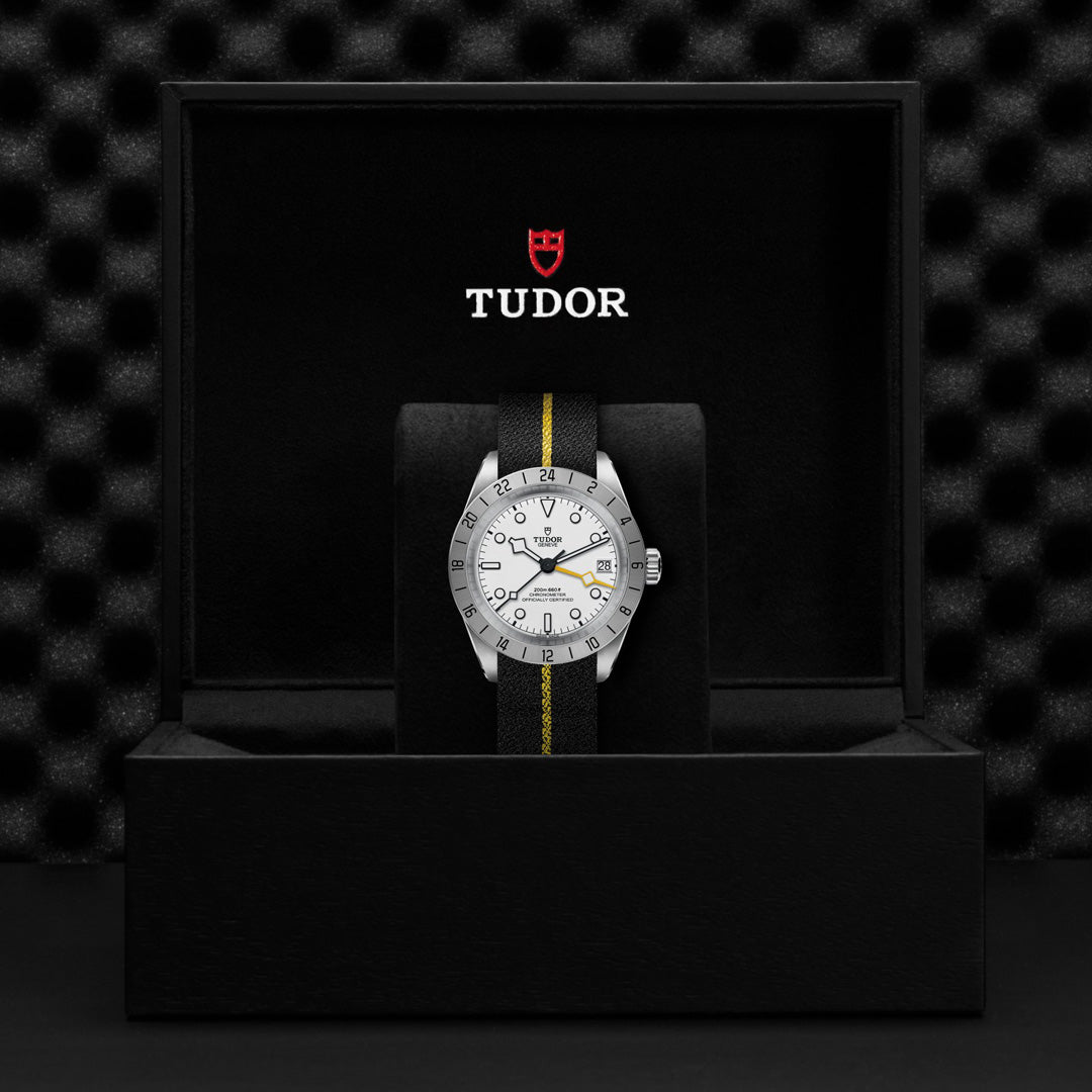 A TUDOR Black Bay Pro watch with a silver bezel, white dial, and black band with a yellow stripe is displayed in an open black box featuring the prominent red TUDOR logo. It houses the TUDOR Calibre MT5652. The backdrop shows a textured black pattern.