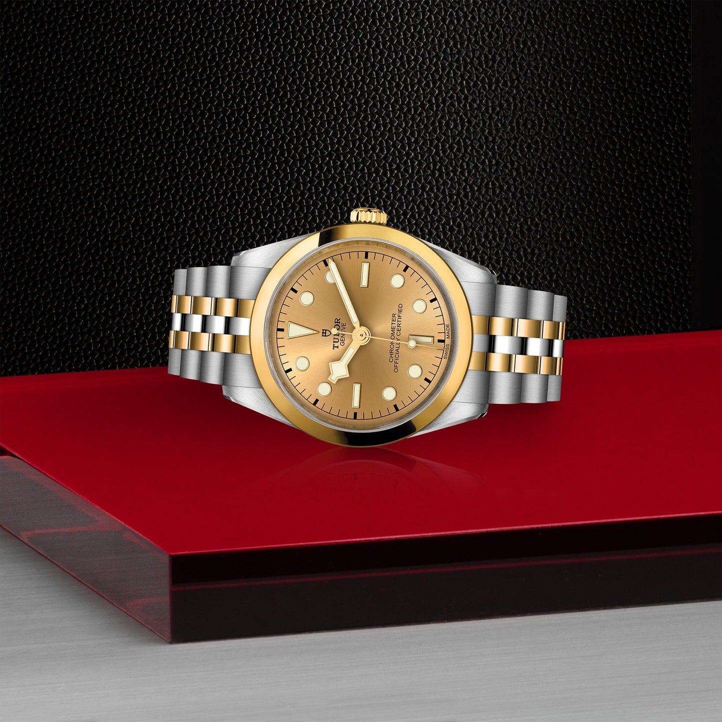 The TUDOR Black Bay One 36 S&G luxury wristwatch rests elegantly on a glossy red surface, featuring a 36mm steel case with a gold dial, stainless steel and gold bracelet, gold hour markers, and a striking black textured background.