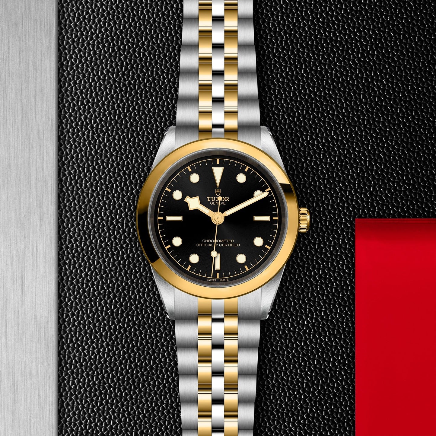 The TUDOR Black Bay One 41 S&G features a 41mm steel case, black dial with gold hour markers and hands, and a two-tone bracelet of gold and silver links. It rests elegantly on a textured black surface with a striking red and silver background.