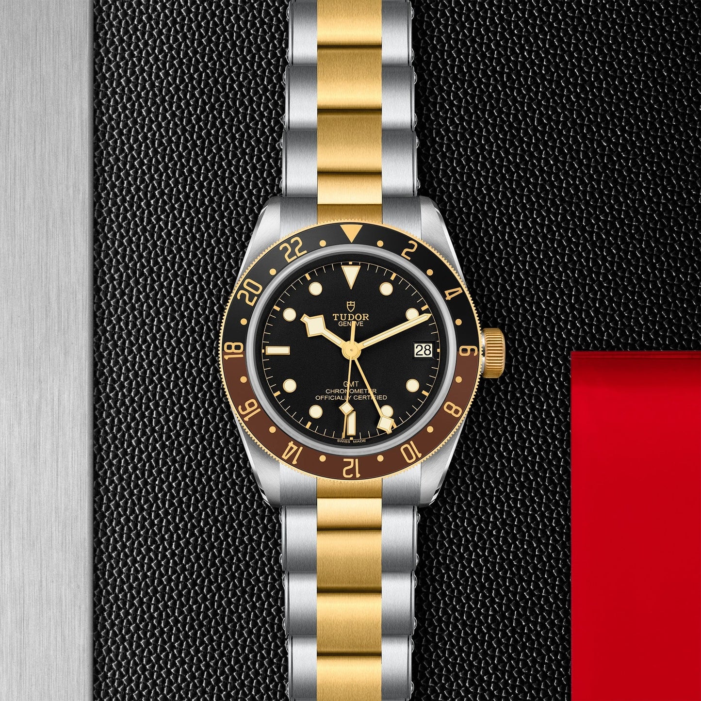 The TUDOR Black Bay GMT S&G wristwatch, featuring a gold and silver metal strap, is placed on a textured black surface. It showcases multiple time-zone features along with a black dial adorned with gold numerals and markers, a date window, and a rotatable bezel. On the right side, part of a small red card can be seen.