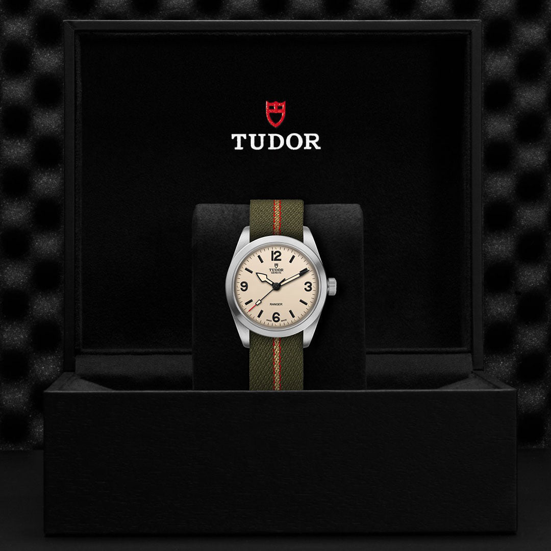 The TUDOR Ranger by TUDOR features a beige dial and green fabric strap, showcased in a black box with the Tudor logo and red shield inside the lid, emphasizing its rugged expedition watch design.