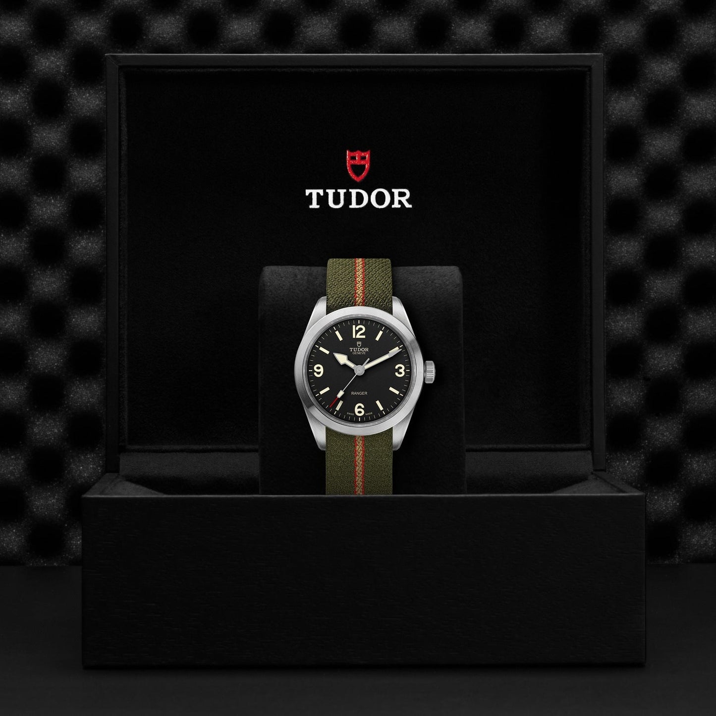 A TUDOR Ranger watch, showcasing a black dial with Arabic numerals, is elegantly presented in an open black box featuring a soft textured interior. This practical timepiece boasts a green and red striped fabric strap, with the TUDOR logo prominently displayed on the box lid.