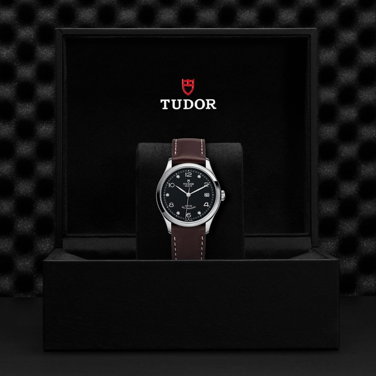 The New Old Stock TUDOR 1926 watch by TUDO1, featuring a diamond-set dial and brown leather strap, is elegantly packaged in a sleek black box. The lid displays the iconic Tudor logo in red and white on a textured black background.