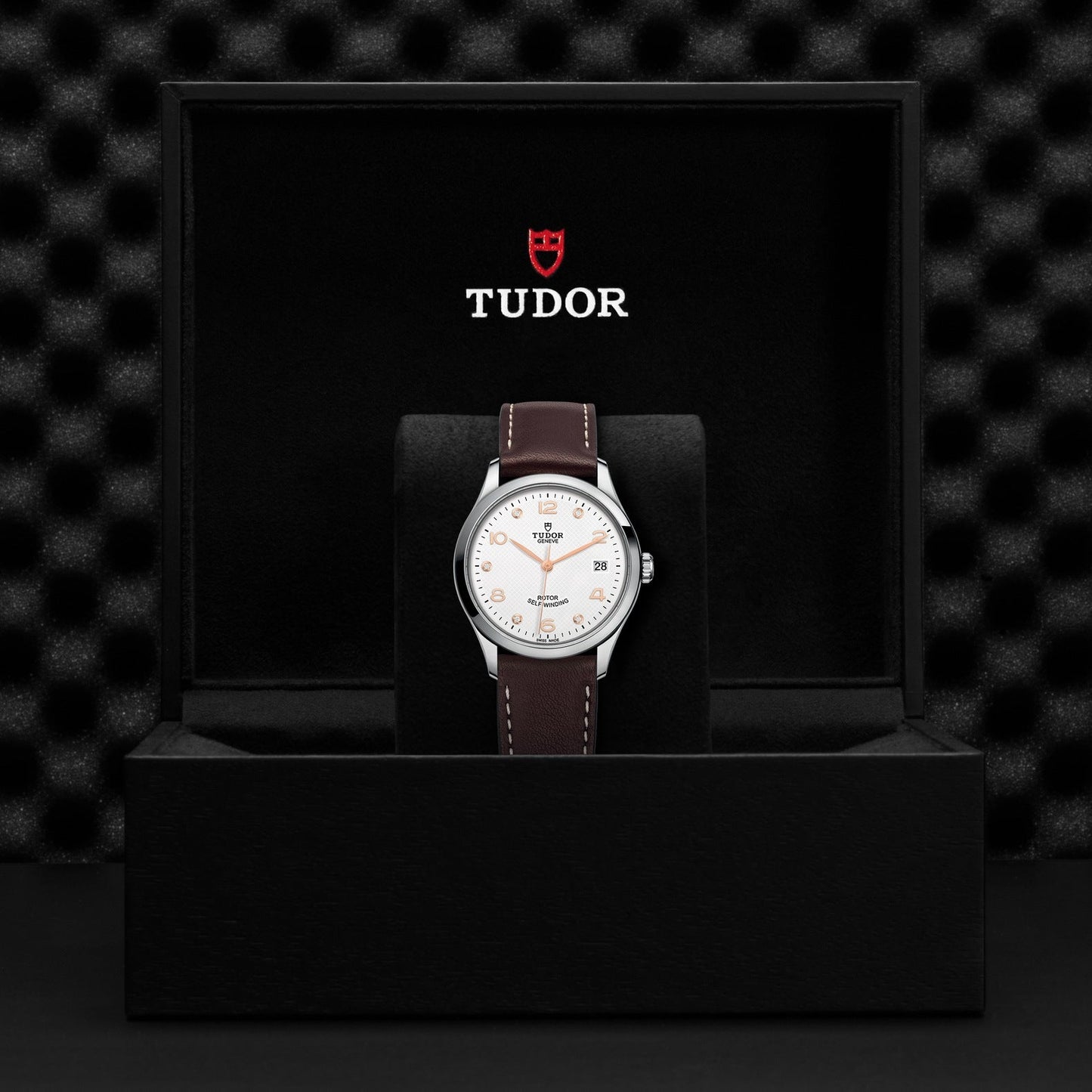 The TUDO1 New Old Stock TUDOR 1926 watch, with a white face and brown leather strap, is elegantly presented in an open black case. The iconic Tudor logo in red and white underscores its craftsmanship against the textured black background, enhancing its timeless allure.