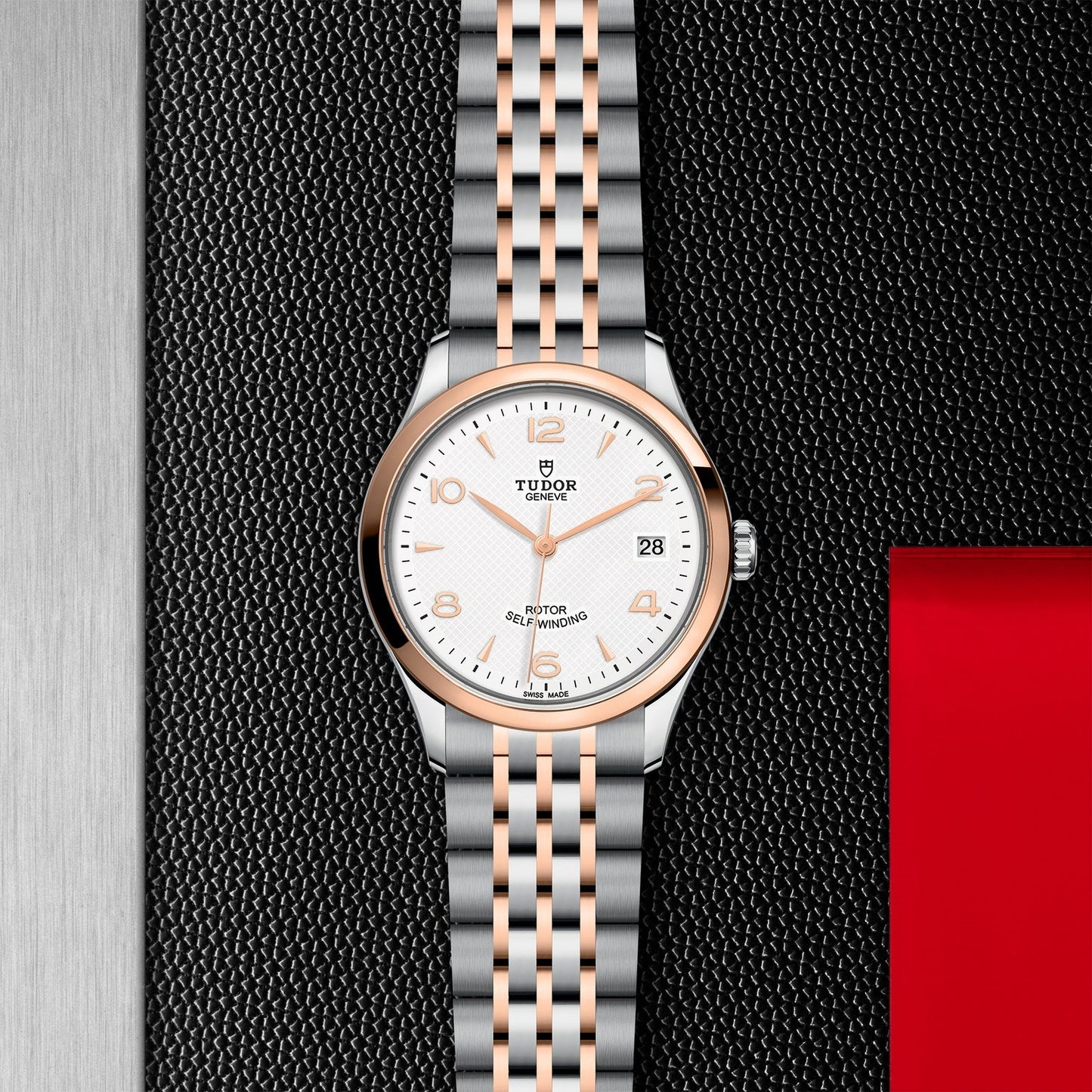 The TUDOR 1926 wristwatch rests against a textured black background with a red accent, showcasing a sophisticated silver and rose gold bracelet. It features a white face adorned with rose gold hour markers, a rose gold bezel, and a date display. The timeless elegance of this piece is accented by the TUDOR branding prominently displayed below the 12 o'clock position.
