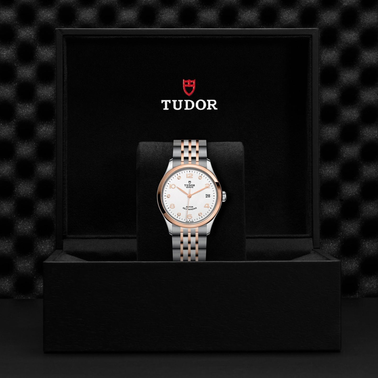 The TUDOR 1926 wristwatch, with its silver and rose gold band, white face, and gold-tone markers, is tastefully presented in an open black box. The brand name TUDOR is prominently featured on the lid of the box.