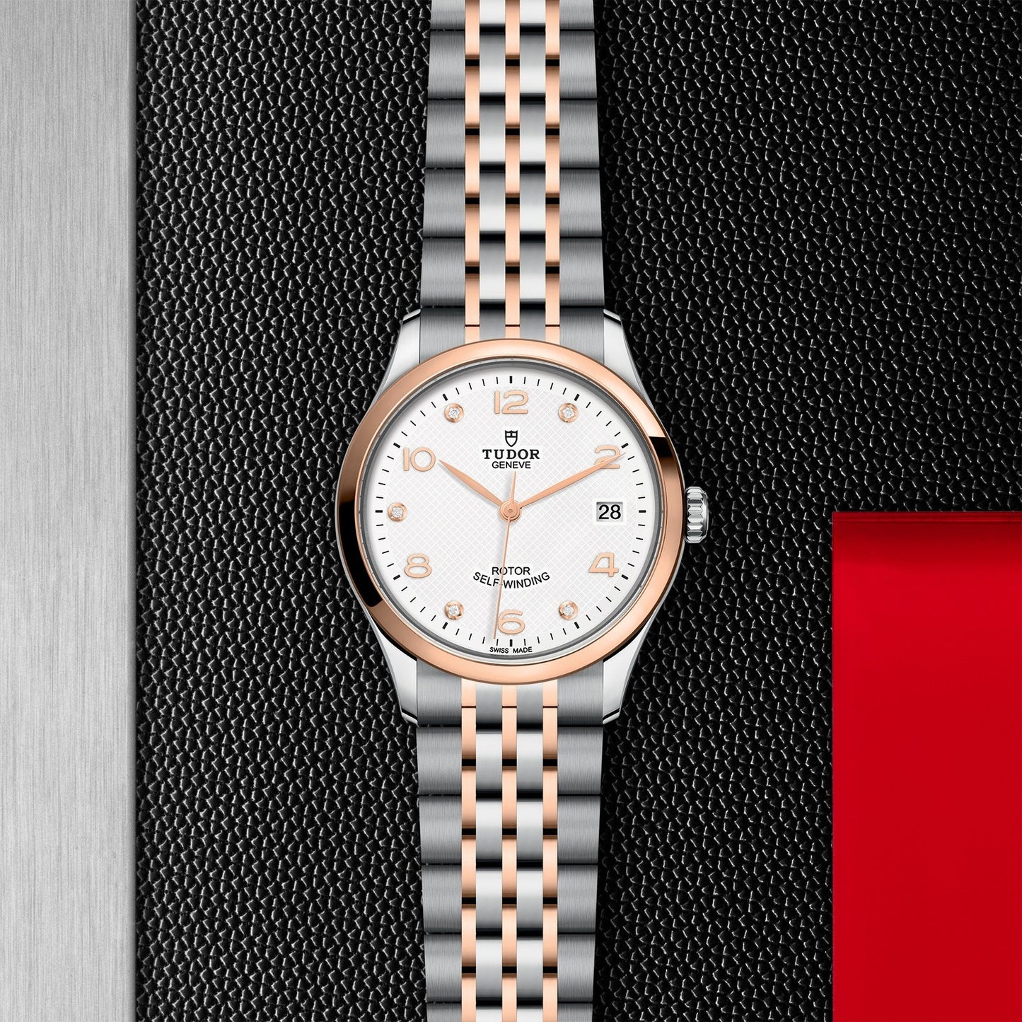 The TUDOR 1926 luxury wristwatch showcases a white dial adorned with gold accents on the hour markers and hands, beautifully paired with a silver and gold two-tone metal bracelet. Set against a textured black and red background, this piece encapsulates the elegance of mechanical watches from the esteemed TUDOR brand.