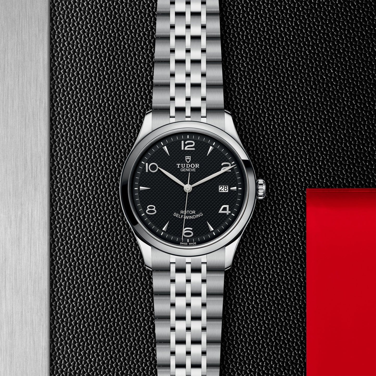 The TUDOR 1926 watch by TUDOR features a black dial, silver hour markers, and a date display. It has a stainless steel bracelet and is showcased on a textured black and silver surface with a touch of red in the corner.