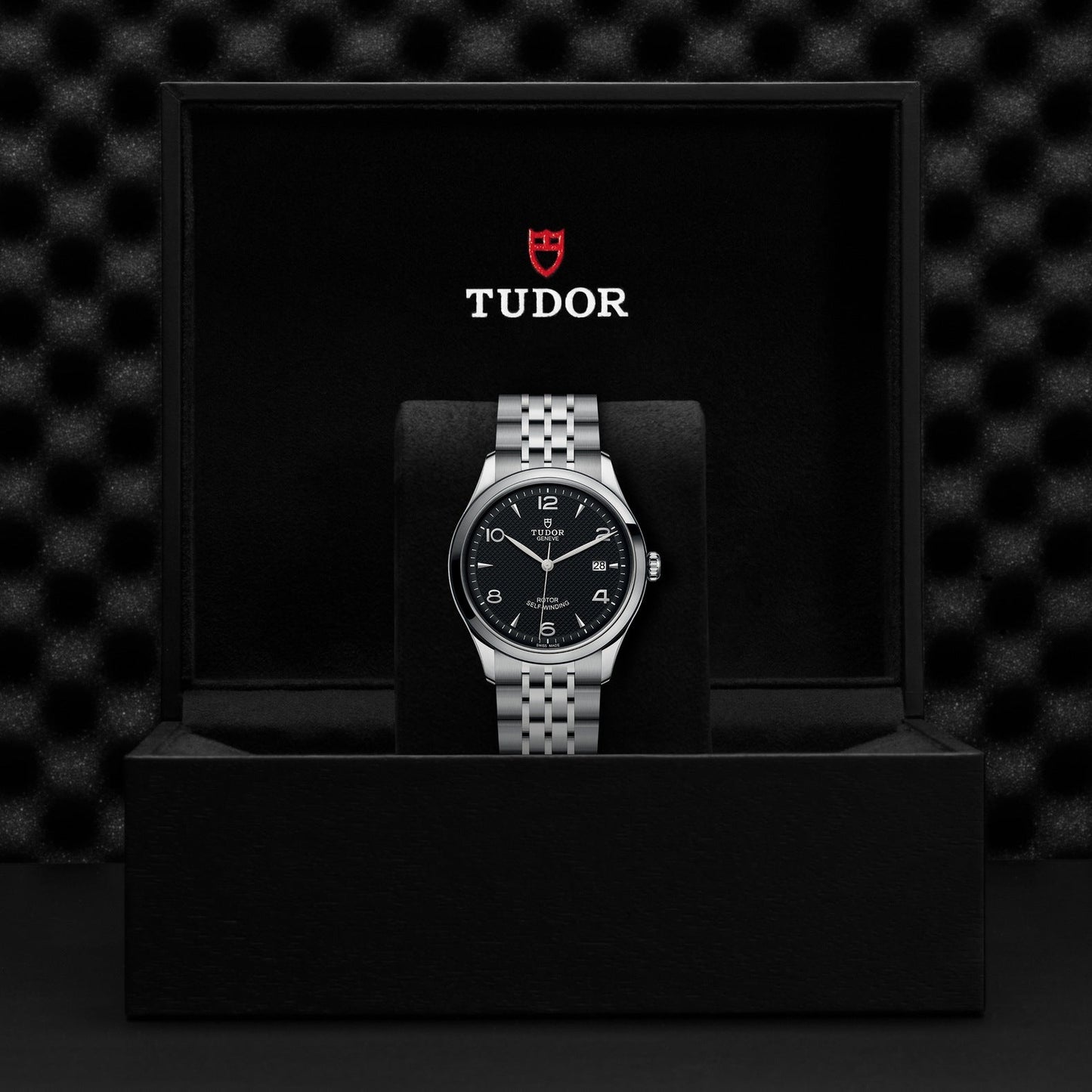 A TUDOR 1926 wristwatch with a black dial and silver metal bracelet is elegantly presented in an open black box, which prominently features the red and white TUDOR logo against a dark, textured background.