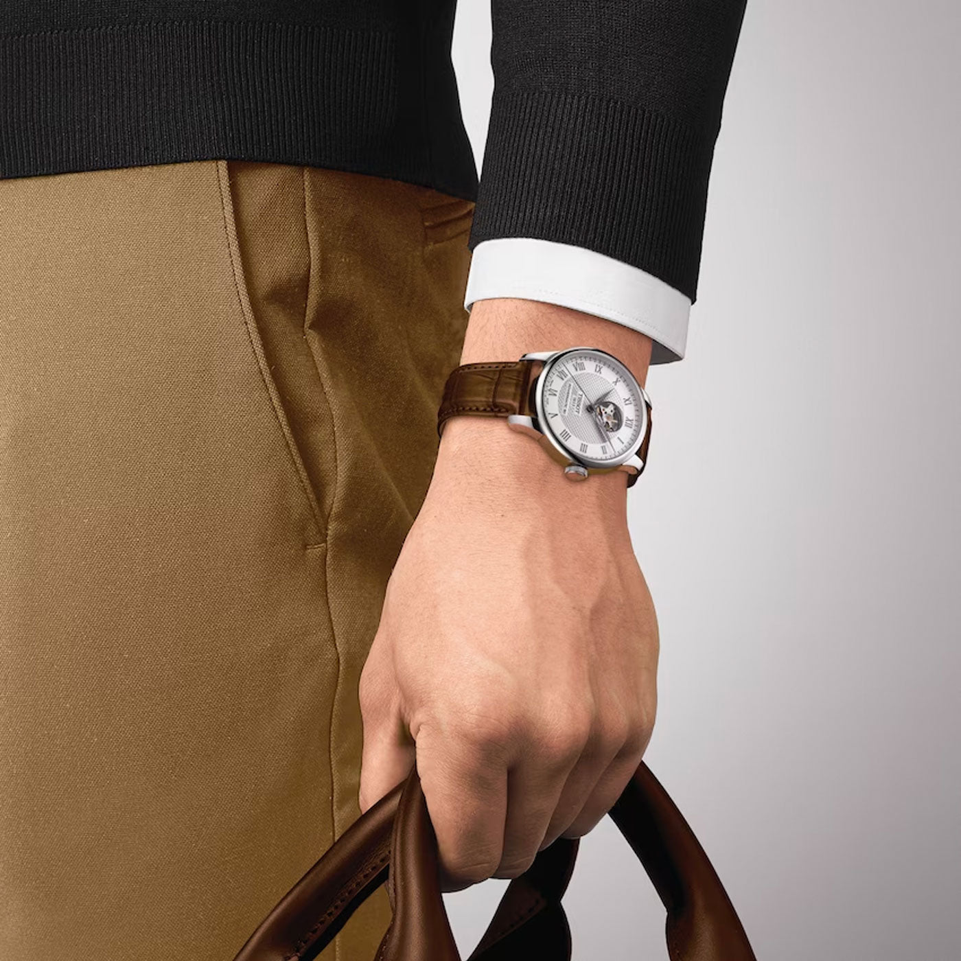 A person is sporting the elegant Tissot Le Locle Powermatic 80 Automatic Open Heart 39.3mm Watch, featuring a brown leather strap and a 316L stainless steel case, with a sophisticated visible mechanical design powered by Swiss automatic movement. They are dressed in brown pants and a black sweater, holding an additional brown leather strap in their other hand.